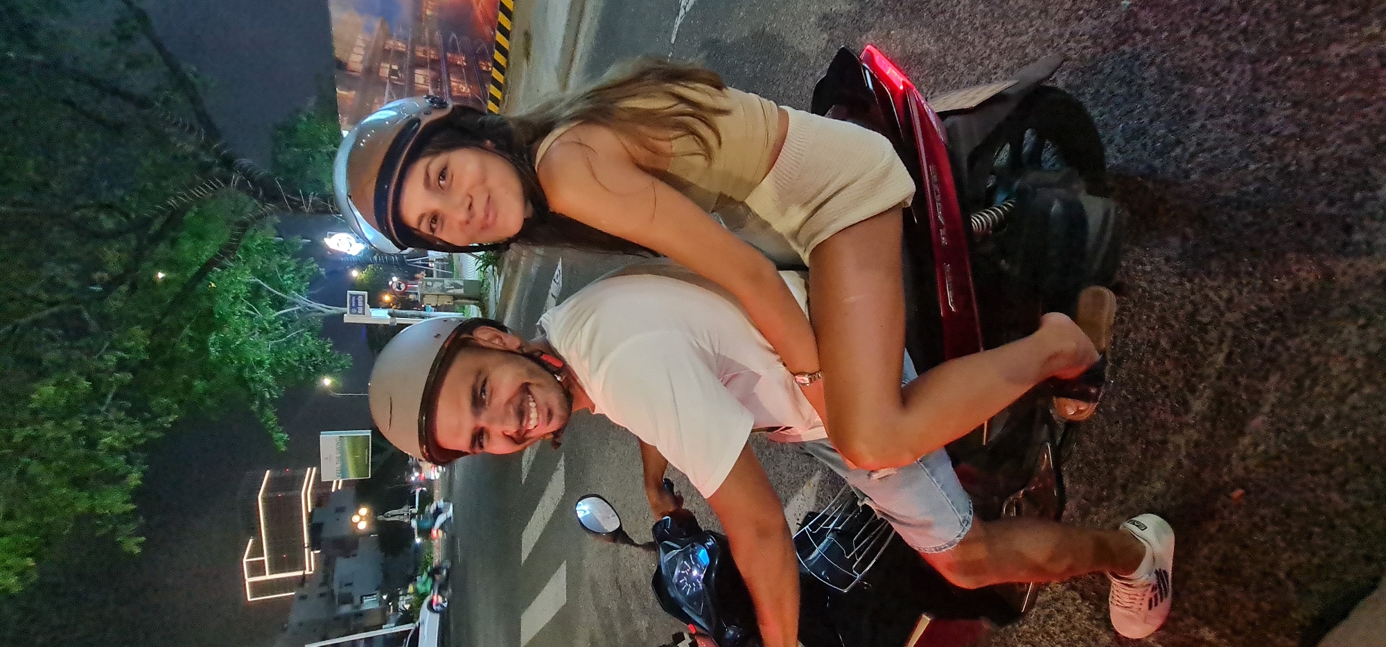 A brightly lit nighttime photograph shows a man and woman riding a motorcycle in Đà Nẵng, Vietnam. Both individuals wear helmets. The man, in a white t-shirt and light blue shorts, is operating the motorcycle and smiling at the camera. The woman, wearing a tan two-piece outfit, sits behind him, also smiling toward the camera. The motorcycle appears to be parked, with the man's right foot on the ground, wearing a white tennis shoe. The woman wears dark slip-on sandals. To the left, trees and city buildings are visible, illuminated by streetlights. The background features signage and architectural details indicative of an urban Vietnamese setting. The road surface appears paved, and the scene is brightly lit, suggesting an active urban area at night.