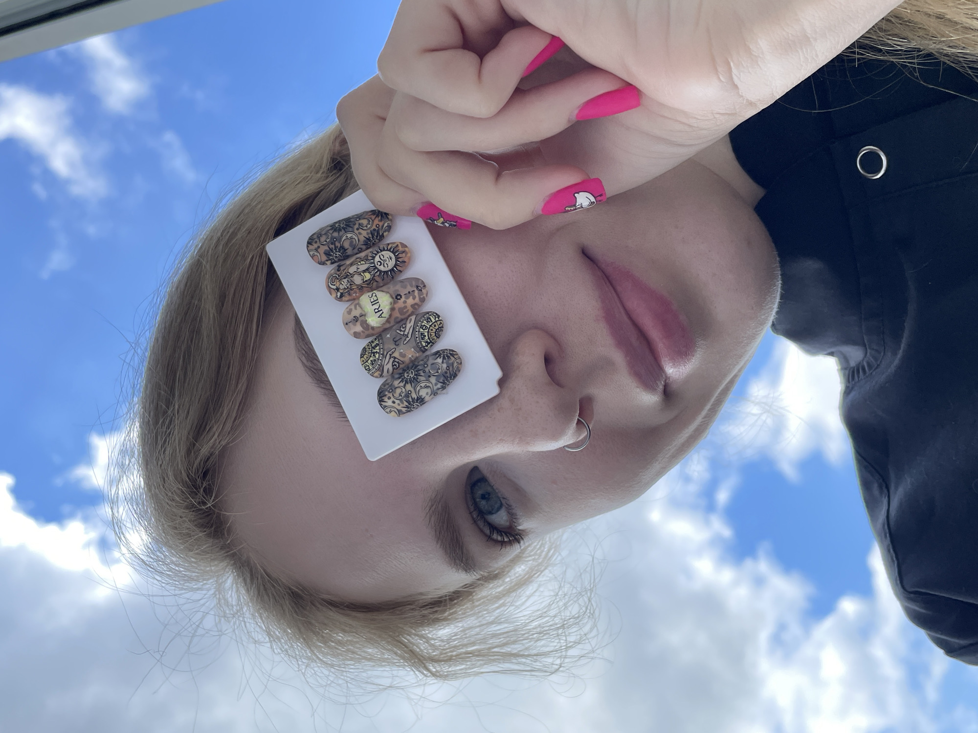 A close-up, low-angle shot, taken in Izhevsk, Russia, captures a young woman showcasing decorated nail tips against a bright blue sky with scattered white clouds. The image is oriented vertically, with the woman's head tilted back, looking towards the viewer.
The woman has blonde hair and a fair complexion. Her left blue eye is visible, framed by light eyebrows and dark eyelashes. A silver hoop nose ring is worn in her left nostril. Her lips are slightly parted, revealing a pink hue. She is dressed in dark, possibly black, clothing, with a silver eyelet visible on the fabric near her shoulder.
Her right hand, with bright pink manicured nails, is held up near her face. The pinky finger's nail features a distinct white cat design, while other nails also bear subtle patterns. She holds a white rectangular card that displays five elongated, artificial nail tips. These tips are intricately adorned with various designs that appear to be astrological or tarot-inspired. One prominent nail tip clearly features the text "Aries," while another shows a stylized sun symbol. The clear sky suggests it is daytime.