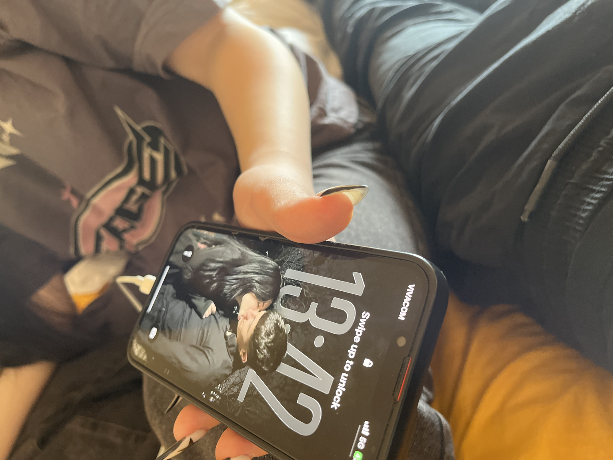 A person is lying down, holding a smartphone in their hand. The phone's screen displays a picture of a couple kissing, along with the time "13:42" and the text "Swipe up to unlock." The phone indicates the carrier is "VIACOM." The person holding the phone is wearing a dark-colored shirt with a logo and has long nails with pointed tips. They also wear dark-colored pants. The background is slightly blurred, showing what appears to be a bed or couch with dark-colored fabric. The location is not indicated by image content. The city of Goce Delcev, Bulgaria is not indicated in image.