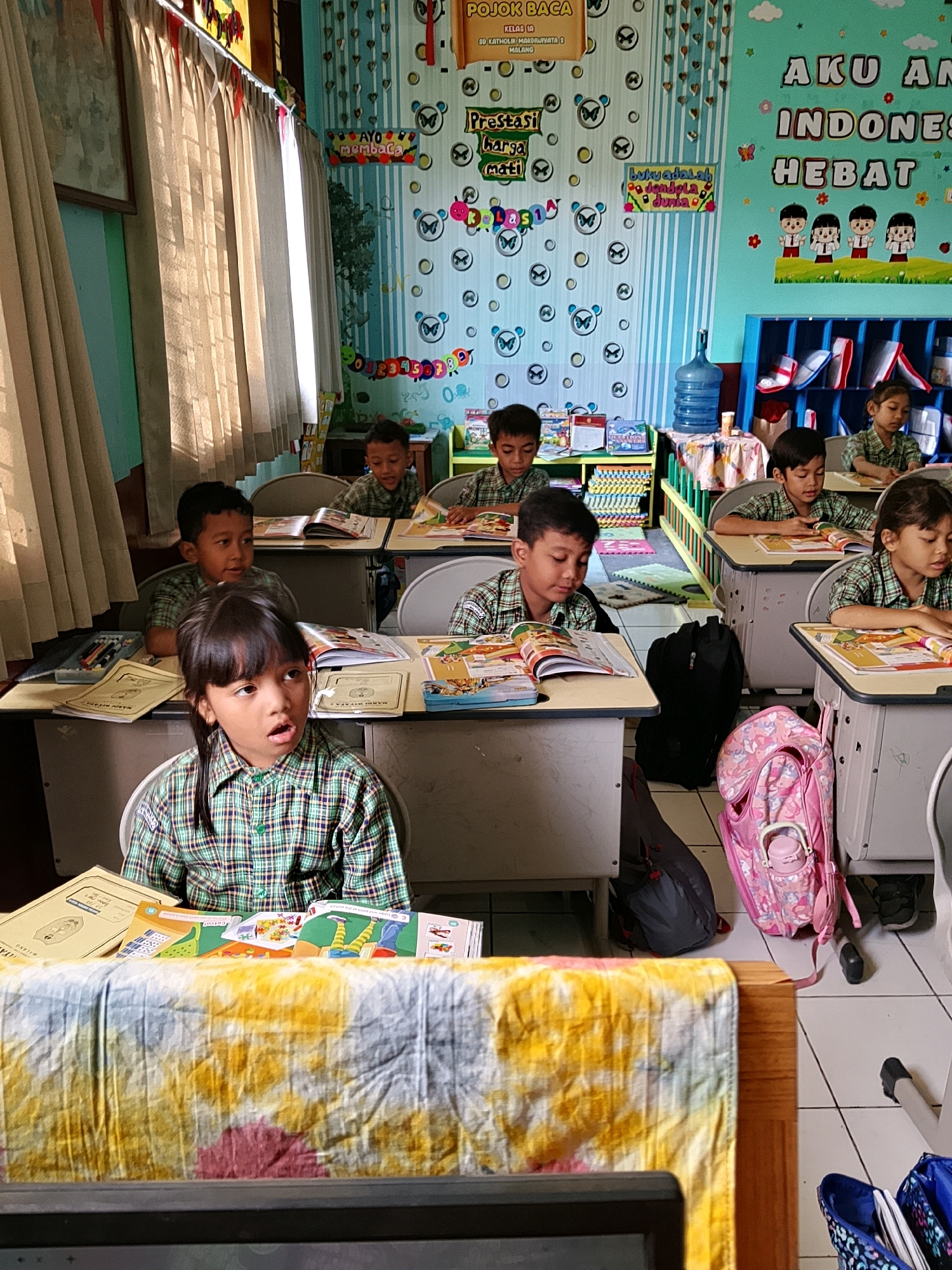 This image depicts a classroom scene at SD Katolik Marsudirini 2 in Malang, Indonesia. Several elementary school students, dressed in plaid shirts, are seated at desks, appearing to be engaged in reading or learning activities. The classroom walls are decorated with colorful posters, including one that reads "AKU ANAK INDONESIA HEBAT" (I am a great Indonesian child) and another with cartoonish characters. Educational materials, such as books and papers, are visible on the desks. Backpacks and a water dispenser are also present in the room. The overall atmosphere suggests a typical school day in a primary educational institution in Indonesia.