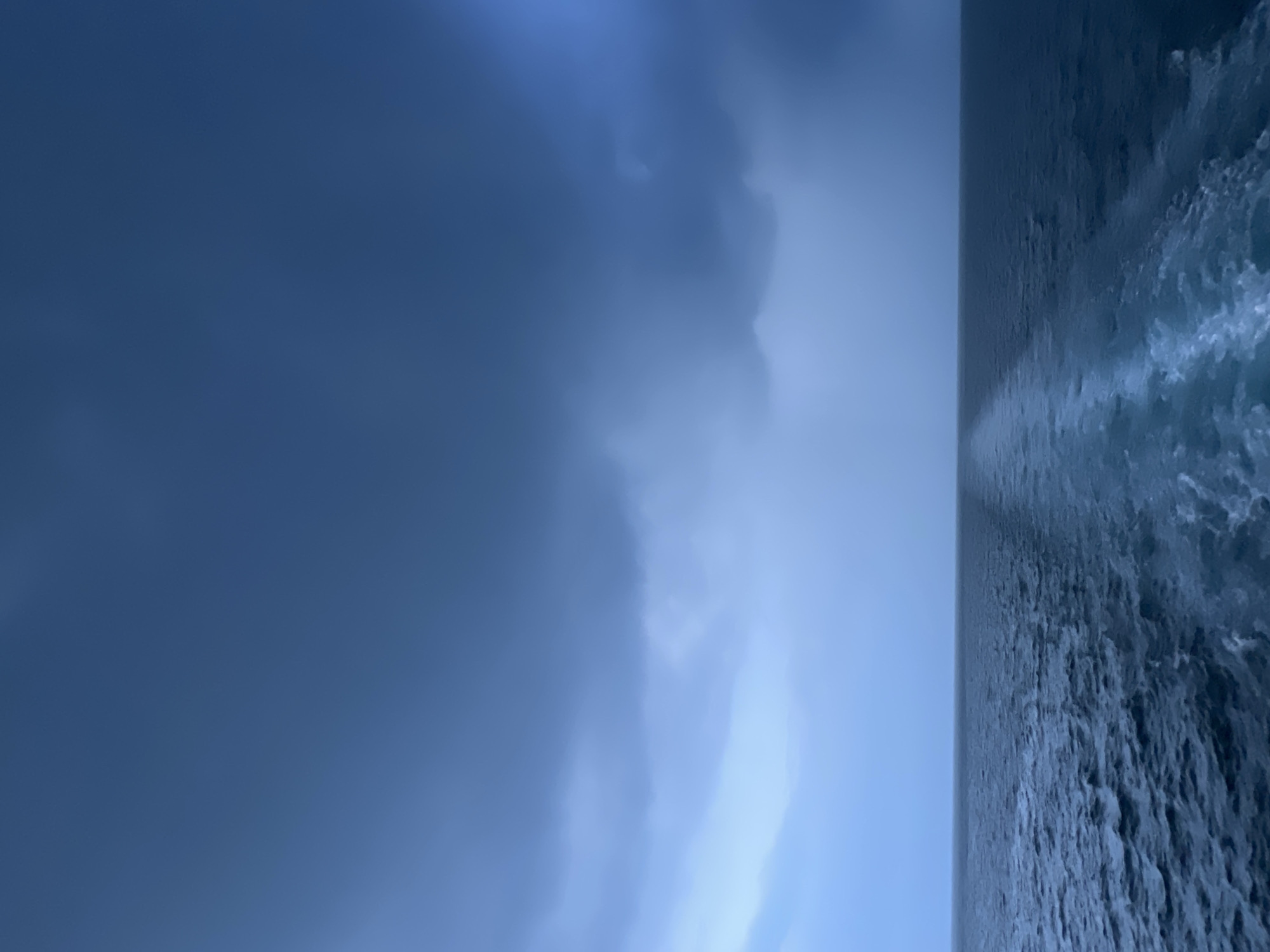 The image depicts a wide expanse of dark blue ocean under a stormy, overcast sky. The horizon is clearly defined, separating the turbulent water from the dramatic cloud formations. A white wake trails behind the unseen vessel from which the photograph is taken, creating a stark contrast against the dark water. The overall atmosphere is somber and foreboding, suggesting the presence of inclement weather. The location is not specified within the city of Unknown, Thailand.
