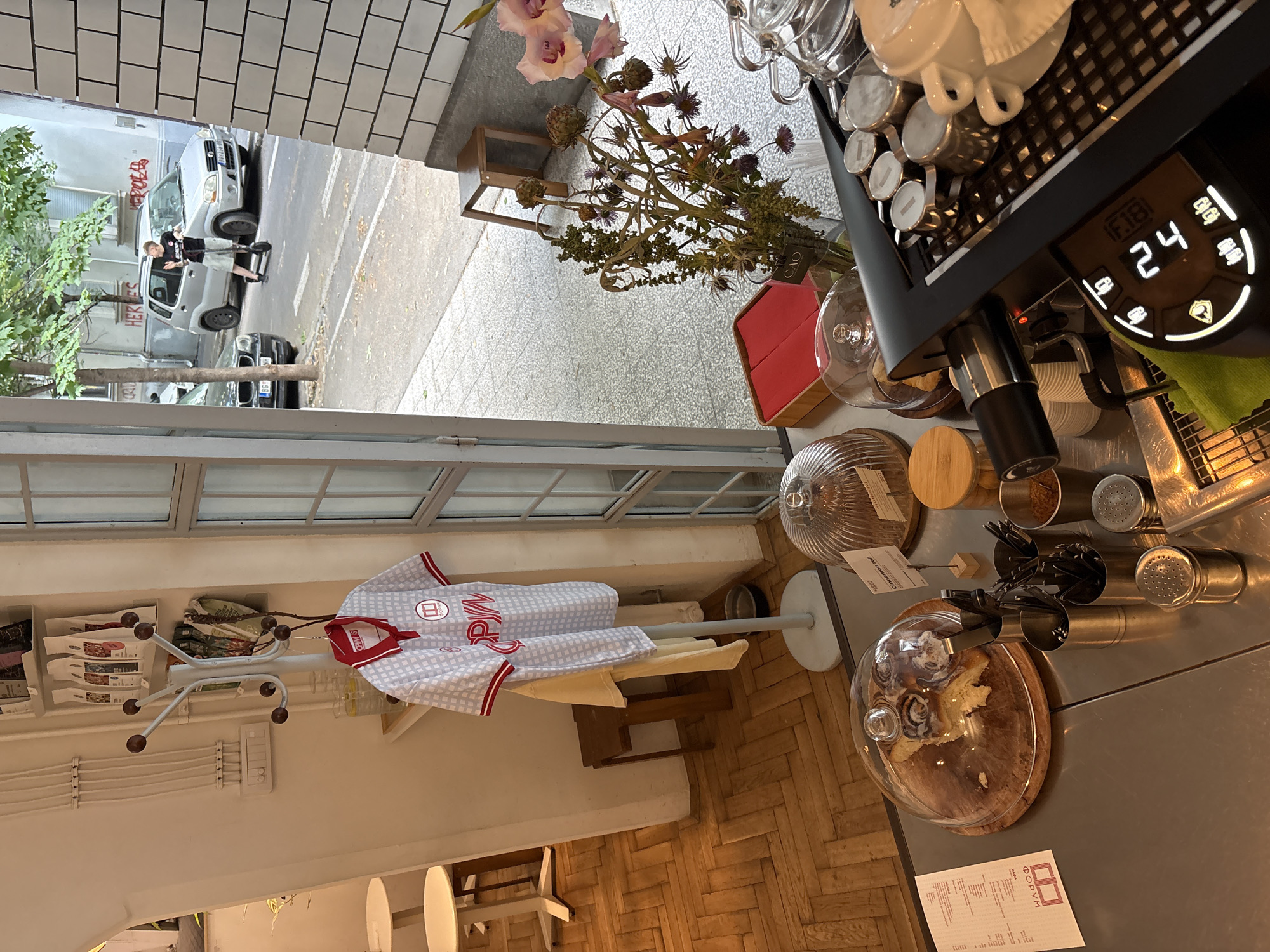 The image shows the interior of a cafe, likely in Belgrade, Serbia, with a view looking outwards to a street scene. Inside, a coat rack holds a patterned shirt. In the foreground, a counter displays cinnamon rolls under glass domes, various utensils, and a coffee machine. To the right, a vase holds dried flowers and a glint of light reflects off a silver kettle. The street scene visible through the window features a person riding an electric scooter, cars, and buildings with graffiti.