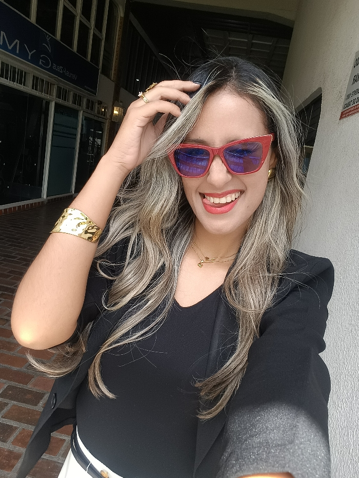 A smiling woman wearing red sunglasses and a black top with a blazer poses outside a building. She is accessorized with a gold bracelet, necklace, and ring. Her blonde hair is styled with dark roots. The background shows a brick walkway, a building facade with glass windows, and a light-colored wall.