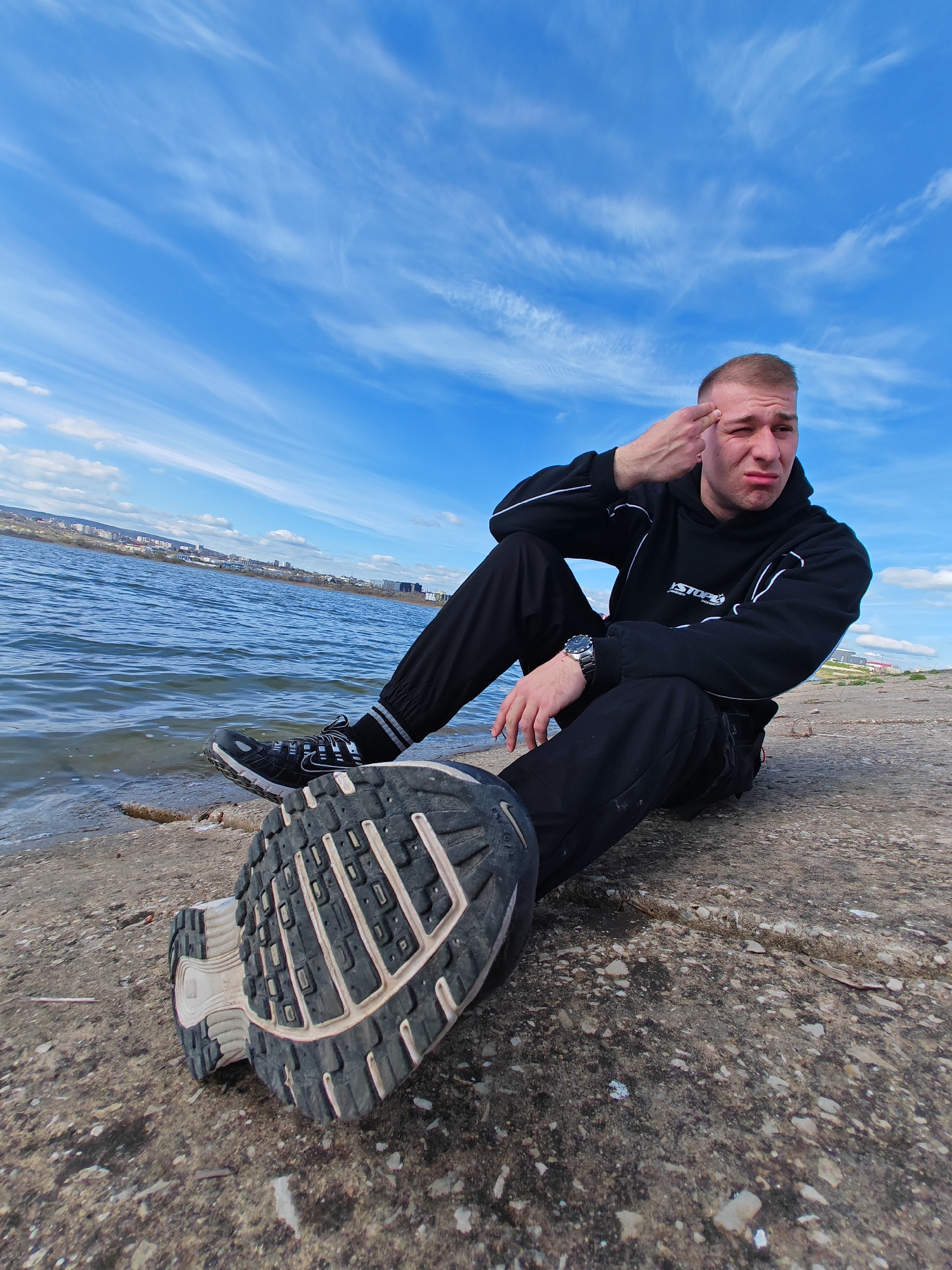 A young man is sitting on a concrete embankment by a body of water. He is wearing a black hooded sweatshirt with white stripes along the sleeves, black pants, and black sneakers with white soles. One of his feet is extended towards the camera, displaying the sole of his sneaker. The man has short blonde hair and is making a gesture with his hand to his forehead. He is also wearing a watch on his left wrist. In the background, across the water, is a cityscape. The sky is blue with streaky clouds.
The location is most likely not in the city of Shumen, Bulgaria. The scene depicts a river or large body of water with a cityscape in the background. Shumen is not located on a major body of water.