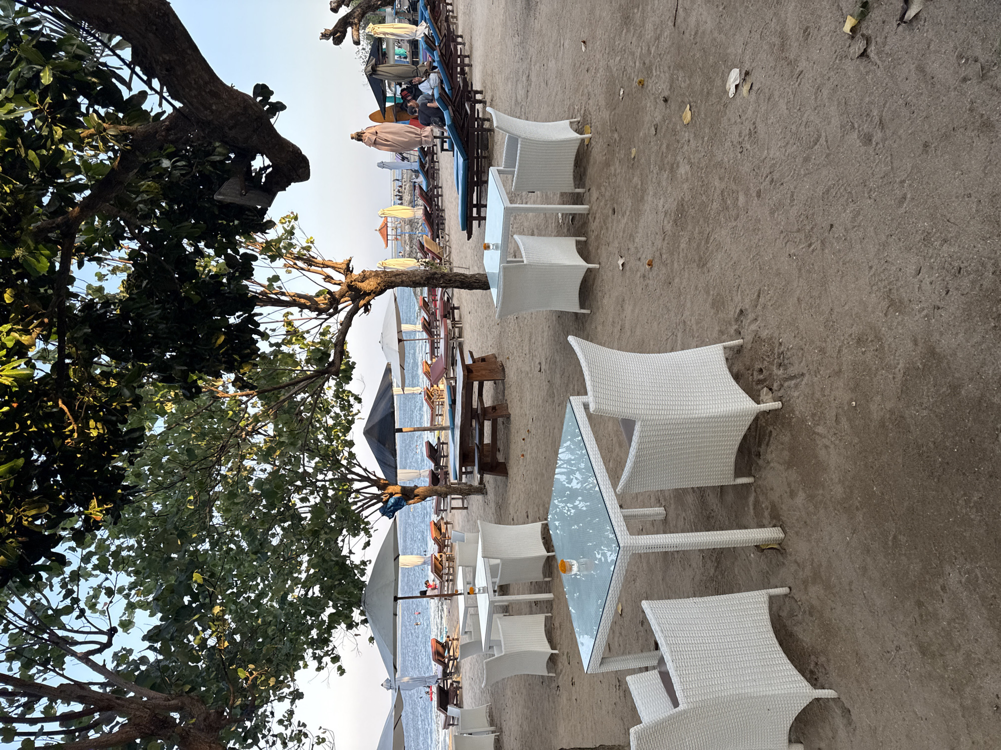 The image depicts a beachside dining area with white wicker furniture and glass-top tables on a sandy beach. In the background, lounge chairs and umbrellas are set up facing the ocean, with a few people visible in the distance. The scene is framed by trees with lush green foliage. The setting appears to be in Denpasar, Indonesia, likely a popular tourist spot for relaxation and dining.