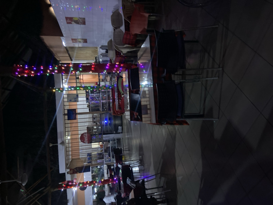 The image captures an outdoor or semi-outdoor dining and gathering area at night, illuminated by numerous colorful string lights. The scene is relatively dim, with artificial lights providing the main illumination.
In the foreground and mid-ground, multiple strands of multi-colored fairy lights are strung horizontally, creating a festive and inviting atmosphere. Below these lights, several tables and chairs are visible on a tiled floor with a diamond pattern, some appearing to be set up for use, others potentially stacked or stored.
In the background, a building or structure with light-colored walls houses what appears to be a commercial establishment, likely a restaurant or cafe. A prominent red circular sign bearing the "Coca-Cola" logo is affixed to one of the walls. Below it, a large glass-fronted refrigerator or display case can be seen, suggesting a beverage or food service area. Other faint posters or decorations are on the walls, but their details and text are not discernible.
A few blurry figures are scattered throughout the space, particularly in the lower left portion of the image, indicating the presence of people. They appear to be engaged in an activity, likely dining or socializing. The time of day is clearly night, evidenced by the darkness and the reliance on artificial lighting for visibility. The overall ambiance is casual and relaxed, fitting for a local eatery or communal space in a location like Bagamoyo, Tanzania.