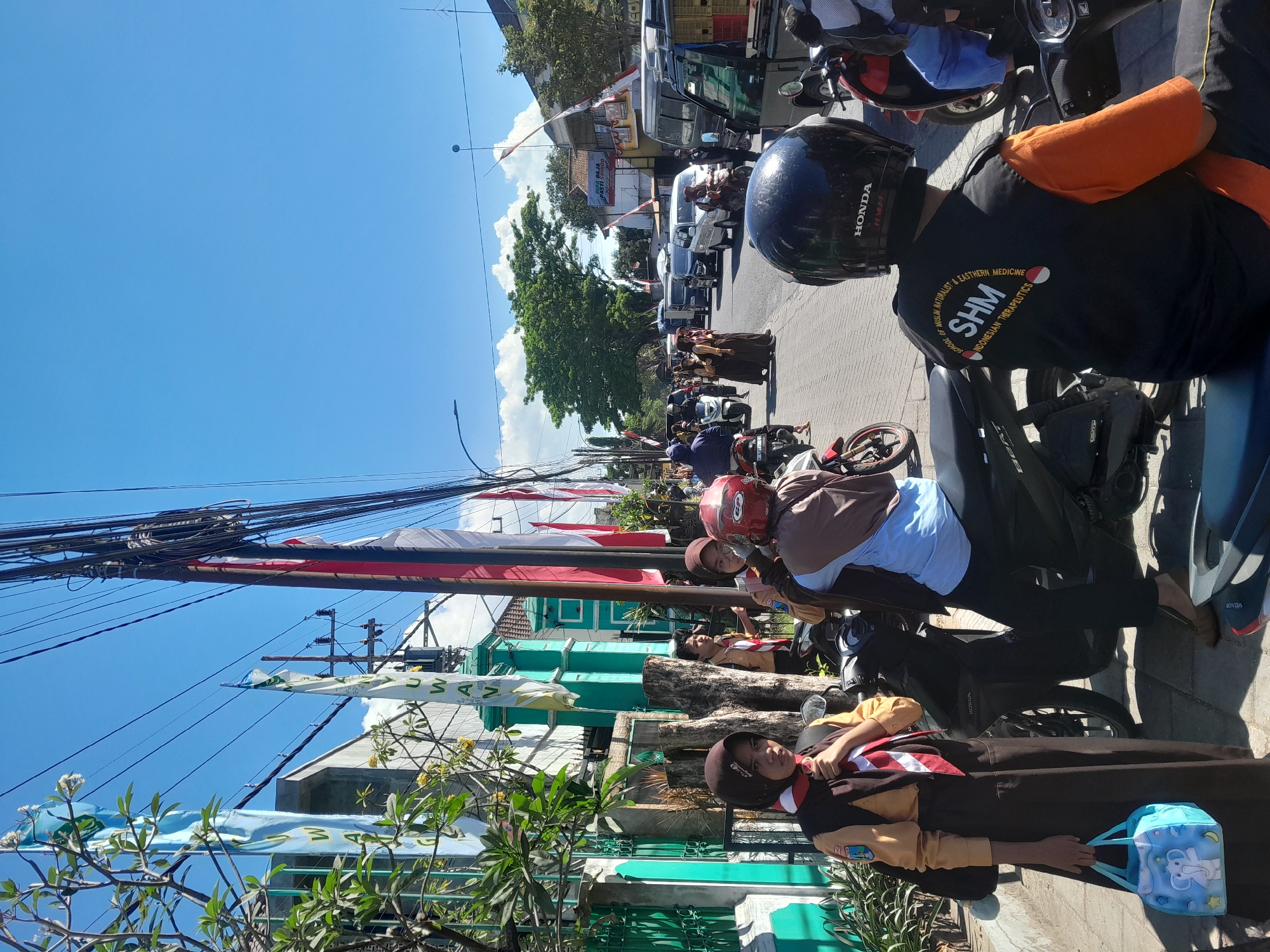 This is a street-level, eye-level, medium shot of a busy street in Srono, Indonesia on a sunny day. In the foreground, a young person wearing a brown uniform with a red and white scarf is standing by a motorcycle. They are holding a light blue lunch bag with an elephant design. To their right, another person wearing a blue shirt and a brown headscarf is seated on a motorcycle. Further down the street, a procession of motorcycles and cars is visible, with some people walking. Red and white flags are prominently displayed along the street, suggesting a cultural event or celebration. The road is paved, and there are buildings and trees lining the sides. Overhead, a dense network of electrical wires is strung across utility poles. The sky is clear blue with a few scattered white clouds.