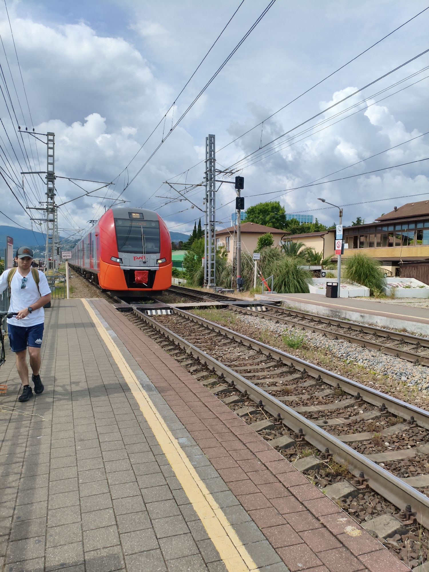 The image depicts a train station platform in Sochi, Russia, on a cloudy day. A person is walking along the platform carrying a bicycle. A red and orange train is approaching on the tracks. The platform is paved with grey tiles and bordered by a yellow line. The train tracks are made of steel rails and wooden sleepers, with gravel ballast. In the background, there are buildings, trees, and power lines. The sky is filled with dramatic, dark clouds, suggesting an impending storm. A sign on the platform is visible, but the text is illegible. The overall scene conveys a sense of travel and the anticipation of arrival or departure.