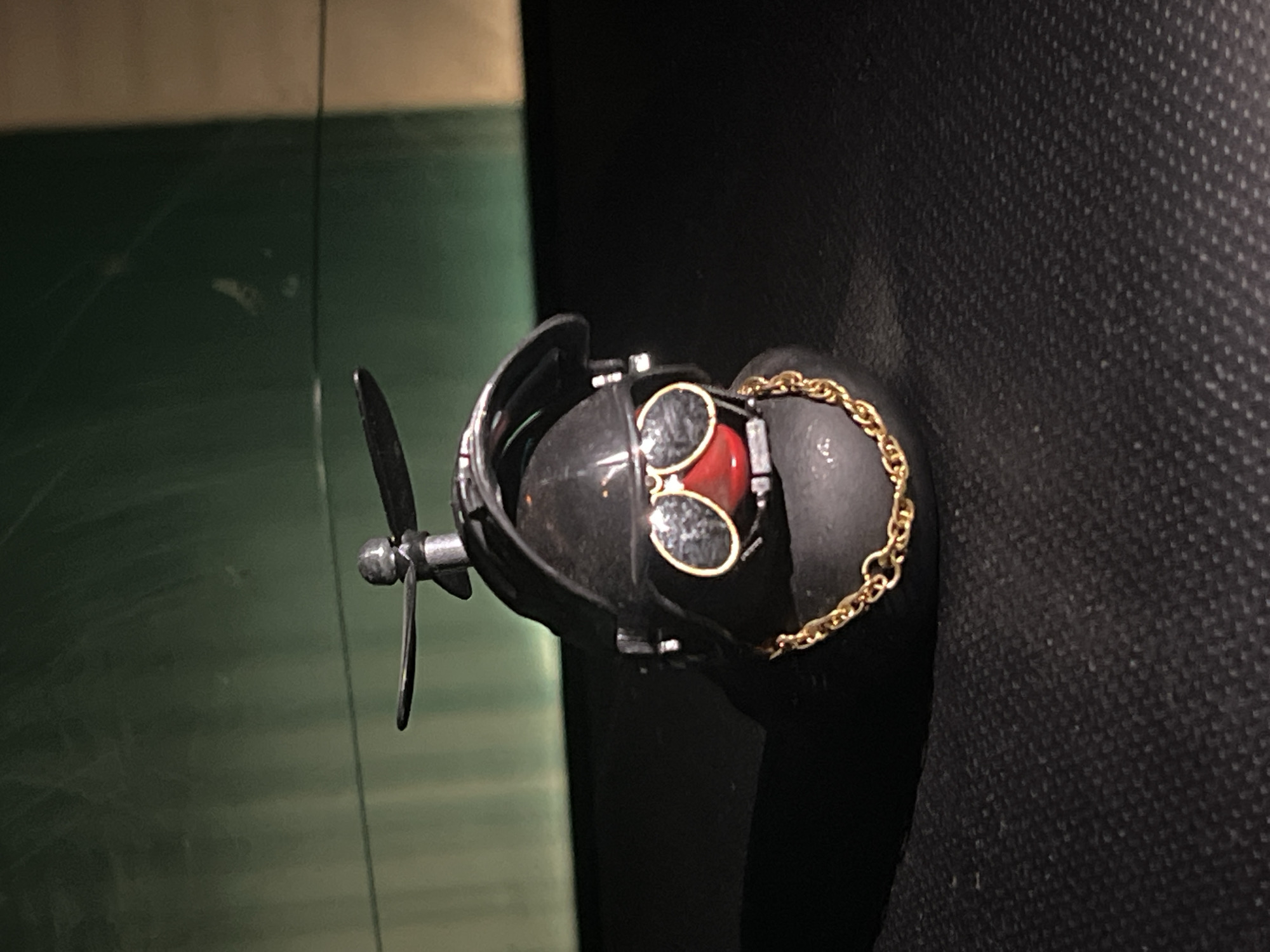 A close-up shows a dark-colored, possibly steampunk-style object featuring a propeller, goggles with red lenses, and a gold chain, resting on what appears to be a dark, textured surface. The object has metallic and black components. A green surface is partially visible in the background. The image doesn't provide information about a location or city.