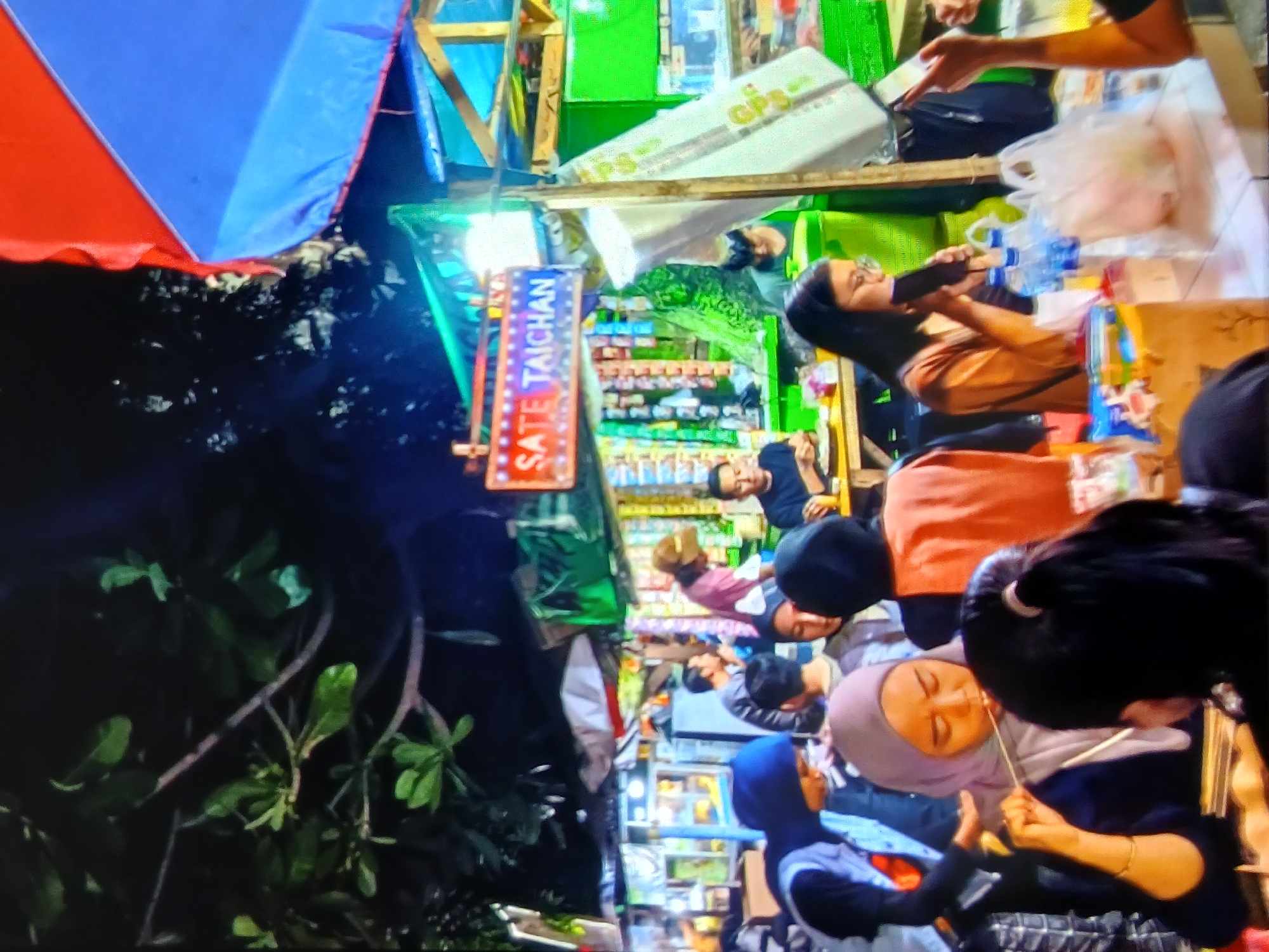 This image depicts a bustling night market scene, likely in Setia Bakti, Indonesia, under a dark sky. A prominent sign reads "SATE TAICHAN," indicating a food stall specializing in this Indonesian satay dish. The stall is decorated with colorful tarps, including red and blue ones overhead, and illuminated by bright lights, casting a vibrant glow on the scene.

Several people are present, engaged in eating and socializing. In the foreground, a woman wearing a light purple hijab is holding a skewer of food, bringing it to her mouth. To her right, another woman with glasses is also holding a skewer and eating. Behind them, other individuals are visible, some seated at tables, suggesting a communal dining experience. The tables are laden with food items, drinks (bottles of water are visible), and plastic bags, typical of a street food environment.

The background shows more stalls and merchandise, hinting at the variety of goods available at the market. The overall atmosphere is lively and energetic, capturing the essence of a local night market.