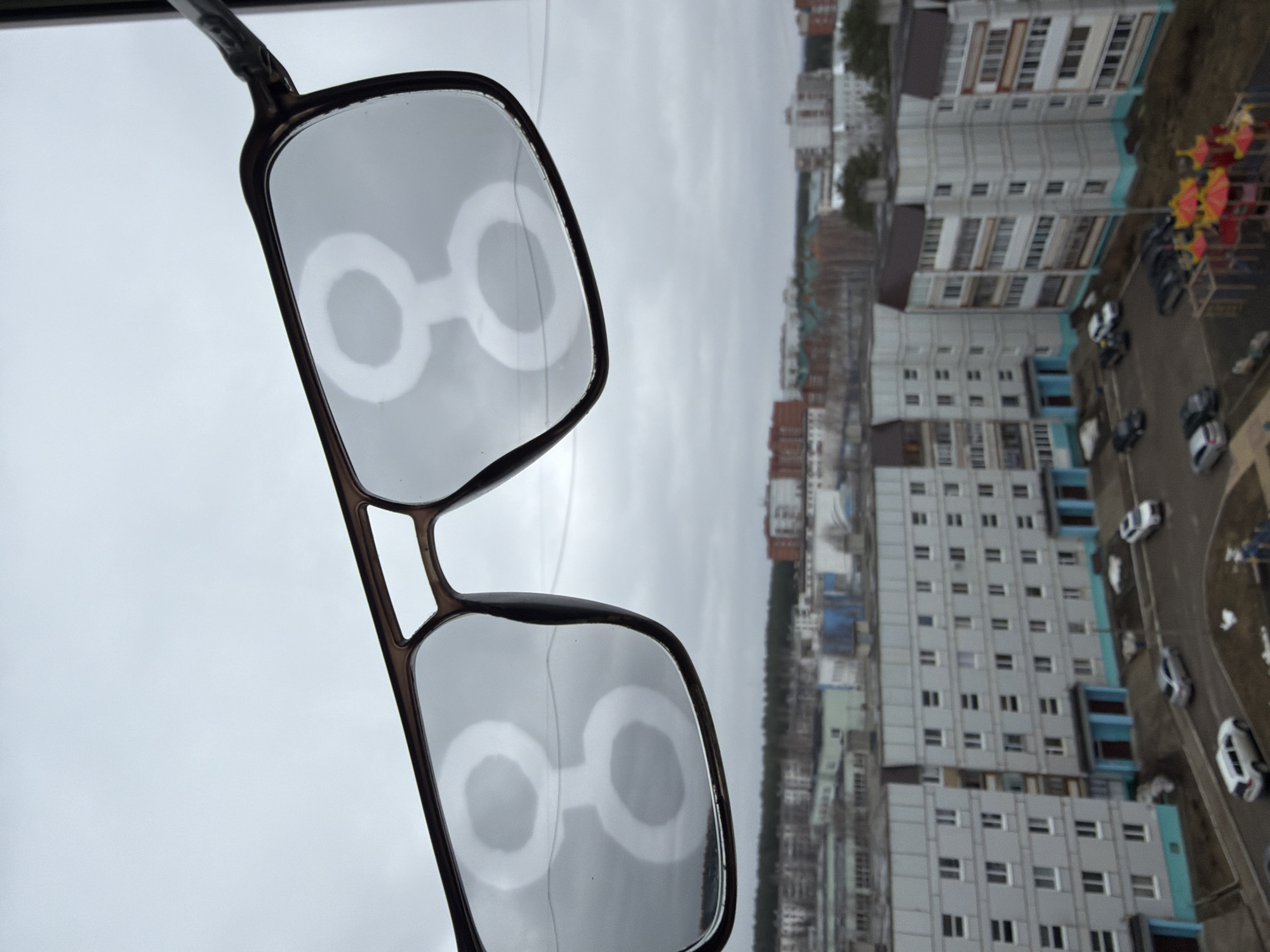 A pair of eyeglasses is held in the foreground against an out-of-focus background featuring multi-story residential buildings in Unknown. The city is overcast. Several cars are visible parked along the side of the road, and there is a playground on the right side. The eyeglasses reflect the same scene.