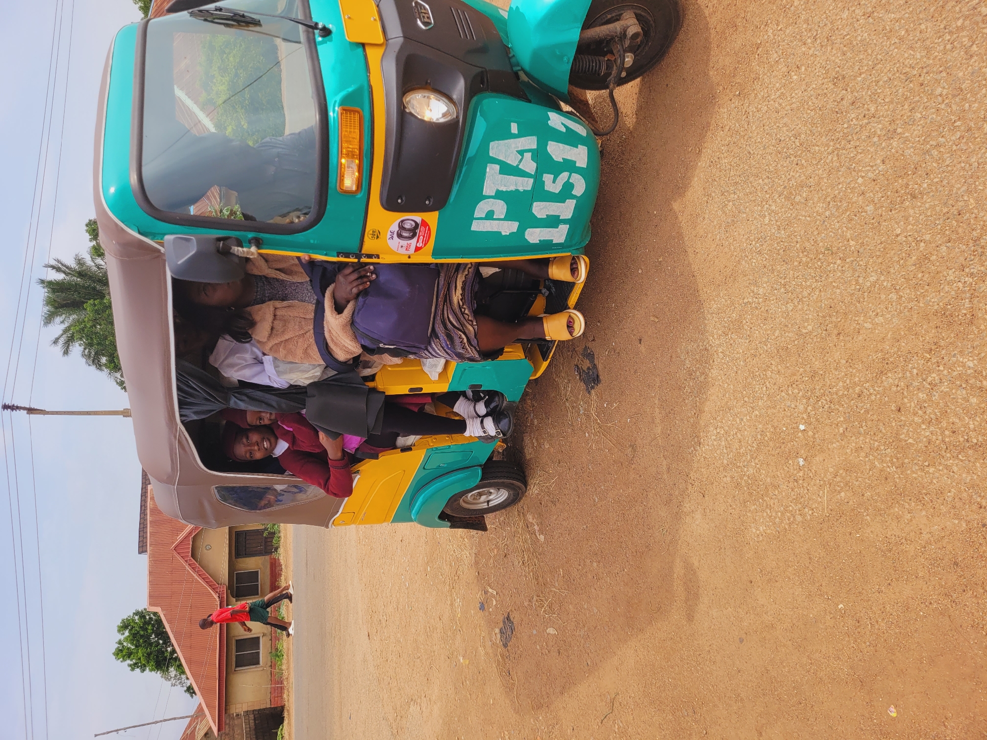 The image depicts a three-wheeled motorized rickshaw, commonly known as an "okada" or "tuk-tuk," with several passengers inside, on a dirt road in Du, Nigeria. The rickshaw is predominantly teal and yellow, with a brown roof. Two young girls, one wearing a maroon sweater and the other in the background partially obscured, are seated inside. A woman in a light brown, furry jacket and a patterned skirt is also a passenger. In the background, a man in a red t-shirt and green shorts walks along the side of the road. To the left of the road, there is a building with a reddish-brown corrugated metal roof. The environment appears to be an arid or semi-arid region, with dusty ground and sparse vegetation. The sky is clear and bright, indicating daytime. The number "PTA-11517" is clearly visible painted on the side of the rickshaw.