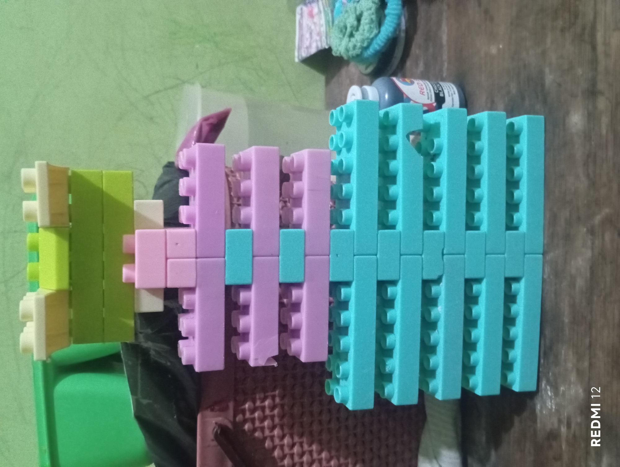 A close-up, slightly tilted shot shows a structure made of interlocking plastic building blocks on a wooden surface. The blocks are stacked in layers, with the bottom layers being light blue, followed by purple layers, and topped with green and white blocks. The background consists of a light green wall and various objects, including a green plastic container, a woven basket, a black plastic bag, and a bottle with a red label. The bottom right corner of the image displays "REDMI 12" watermark. The scene does not provide any specific information about the location in Pagar Alam, Indonesia.