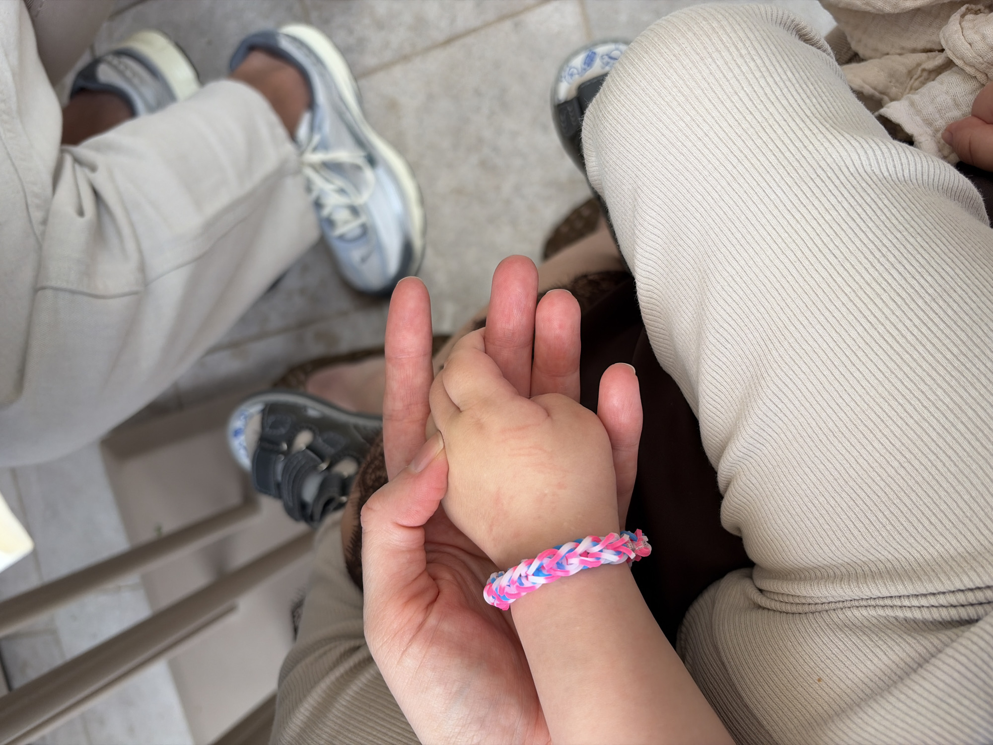 A view from above captures a fair-skinned adult holding the hand of a young child. The child wears a bracelet made of pink, white, and blue rubber bands. The adult's hand encompasses the child's, and the scene includes portions of two people sitting: an adult with silver shoes, and an individual wearing a tan-colored knit material and black and grey sandals. The background includes grey textured flooring, possibly tile or concrete. Parts of a slatted chair can be seen in the lower left. While the exact location in Đenovići, Montenegro is not ascertainable from the image, this image likely has been taken in an indoor environment.