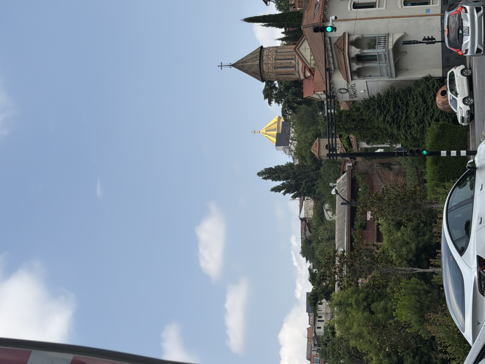 In Tbilisi, Georgia, on a cloudy day, an outdoor view captures a street scene with traffic and a backdrop of buildings and greenery. The foreground is dominated by the rear of a white Tesla Model 3, its sleek design contrasting with the urban environment. To its right, a silver Honda Fit with a license plate starting with "BA" is parked.
Further into the scene, a white SUV is parked on the left, next to a large, distinctive earthenware vessel, likely a qvevri, a traditional Georgian wine vessel. Across the street, a building with the sign "VINOTEL" is visible, featuring traditional Georgian architectural elements like a balcony with intricate metalwork.
The middle ground is a lush expanse of trees and foliage, covering a hillside. Several buildings, some with red-tiled roofs, are nestled among the trees, suggesting a residential area. Dominating the skyline are two prominent religious structures: a church with a conical, stone-covered tower topped with a cross, and behind it, a golden-domed building, possibly a cathedral, gleaming in the daylight. Traffic lights show green, indicating the flow of vehicles. The sky is a soft blue, dotted with scattered white clouds.