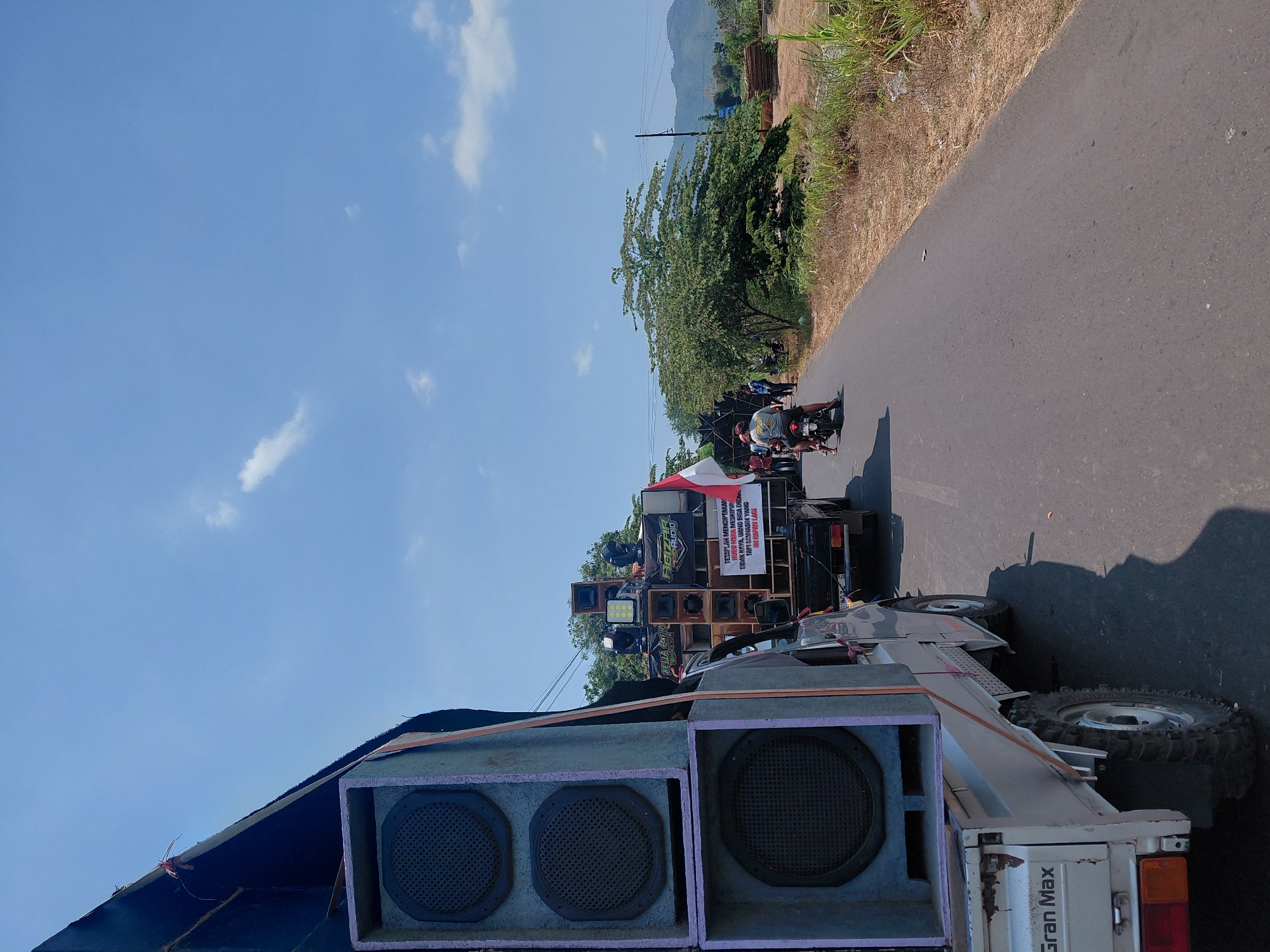 In Talkandang, Indonesia, a procession moves along a paved road under a clear blue sky with scattered white clouds. The foreground is dominated by the side of a white pickup truck, identified by "Gran Max" lettering, loaded with large, stacked speakers. These speakers are housed in purple-edged boxes. In the middle ground, another truck, laden with an array of speakers and sound equipment, is visible. A large Indonesian flag, red and white, is mounted on this truck. Several people are present, some standing near the speaker truck, and one person rides a motorcycle in the lane ahead, followed by others. The roadside features dry, tall grass and vegetation, with trees and distant hills forming the background. The scene suggests a mobile sound system or a parade.