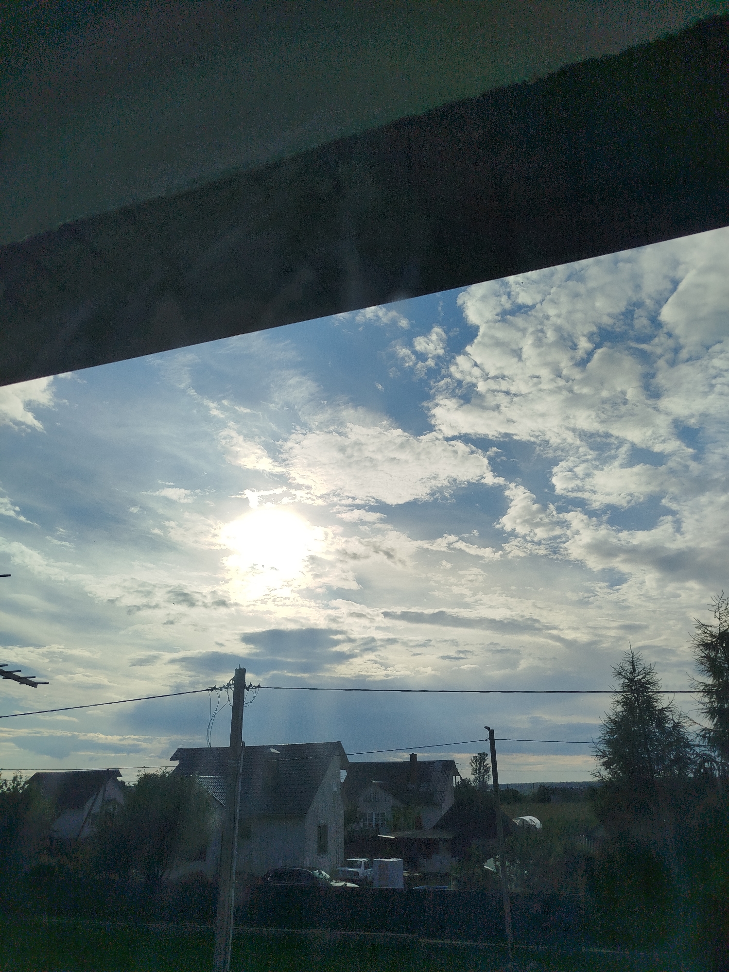 The image depicts a view from an interior space looking out at a cloudy sky over a rural landscape. The sky is a mix of blue and white clouds, with the sun prominently visible, partially obscured by clouds, casting a bright glow. Below the sky, a dark, horizontal structure, possibly an awning or window frame, occupies the top portion of the image.
The landscape below features several houses with pitched roofs, surrounded by trees and vegetation. Utility poles with electrical wires are visible, suggesting a populated area. There are also cars parked near the houses. The overall scene appears to be a residential area in a rural or suburban setting. The location provided, Крывасельскі сельскі Савет, Belarus, indicates a specific rural administrative division within Belarus. The time of day appears to be daytime, possibly late afternoon or early evening, given the angle of the sun.