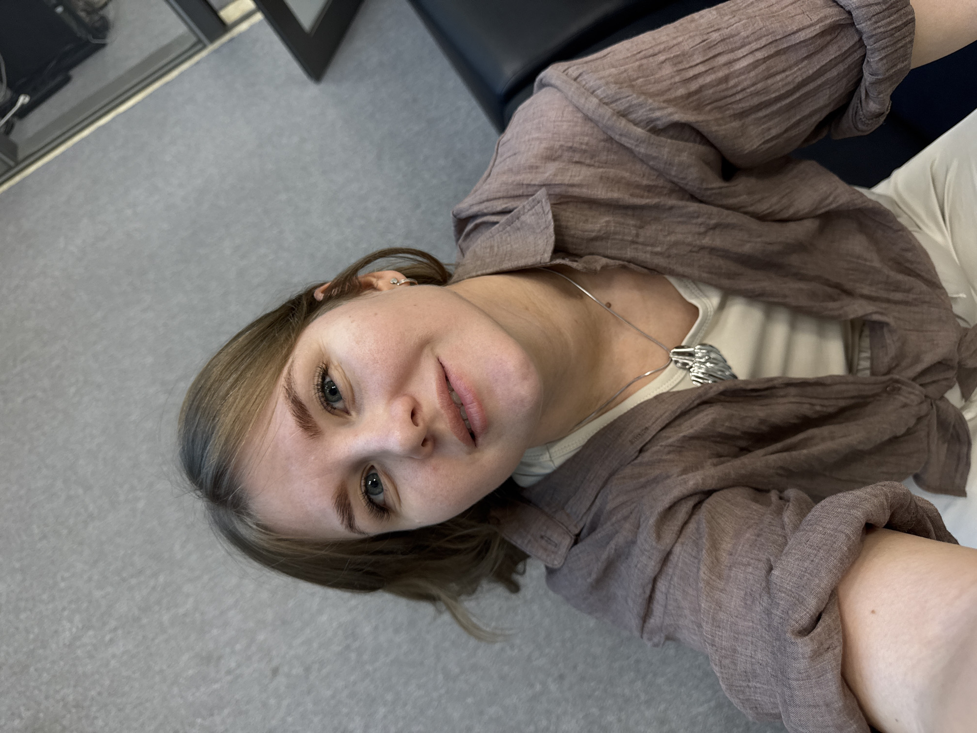 A person is photographed from above, in an indoor location that appears to be in Perm, Russia. The person has fair skin, light brown hair with blonde highlights, and blue eyes. They are wearing a light-colored t-shirt, a brown, open-front, lightweight shirt, and a silver necklace. The person is sitting on a dark chair, and there is grey flooring in the foreground. A window with a black frame is in the top left corner, and a blurry object is reflected in it.