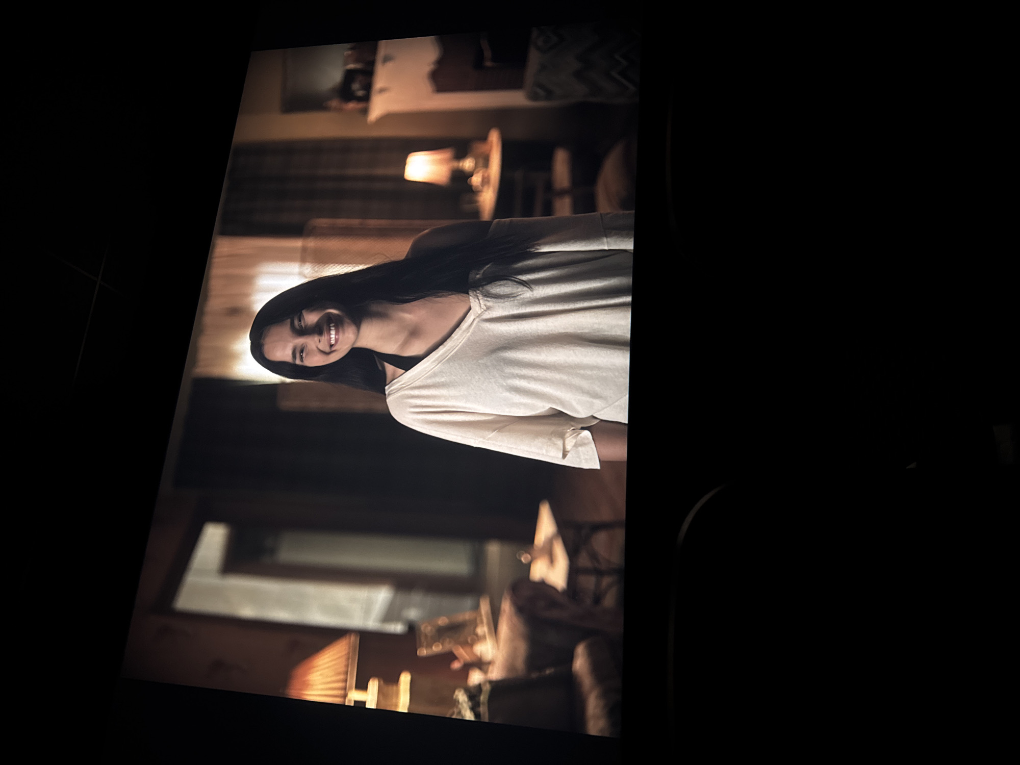 A slightly rotated indoor scene shows a woman with long, dark hair, wearing a white t-shirt and smiling. She appears to be lying down in a room with a bed, a lamp, and other furnishings visible in the background. There is no indication that the image was taken in Puebla, Mexico.