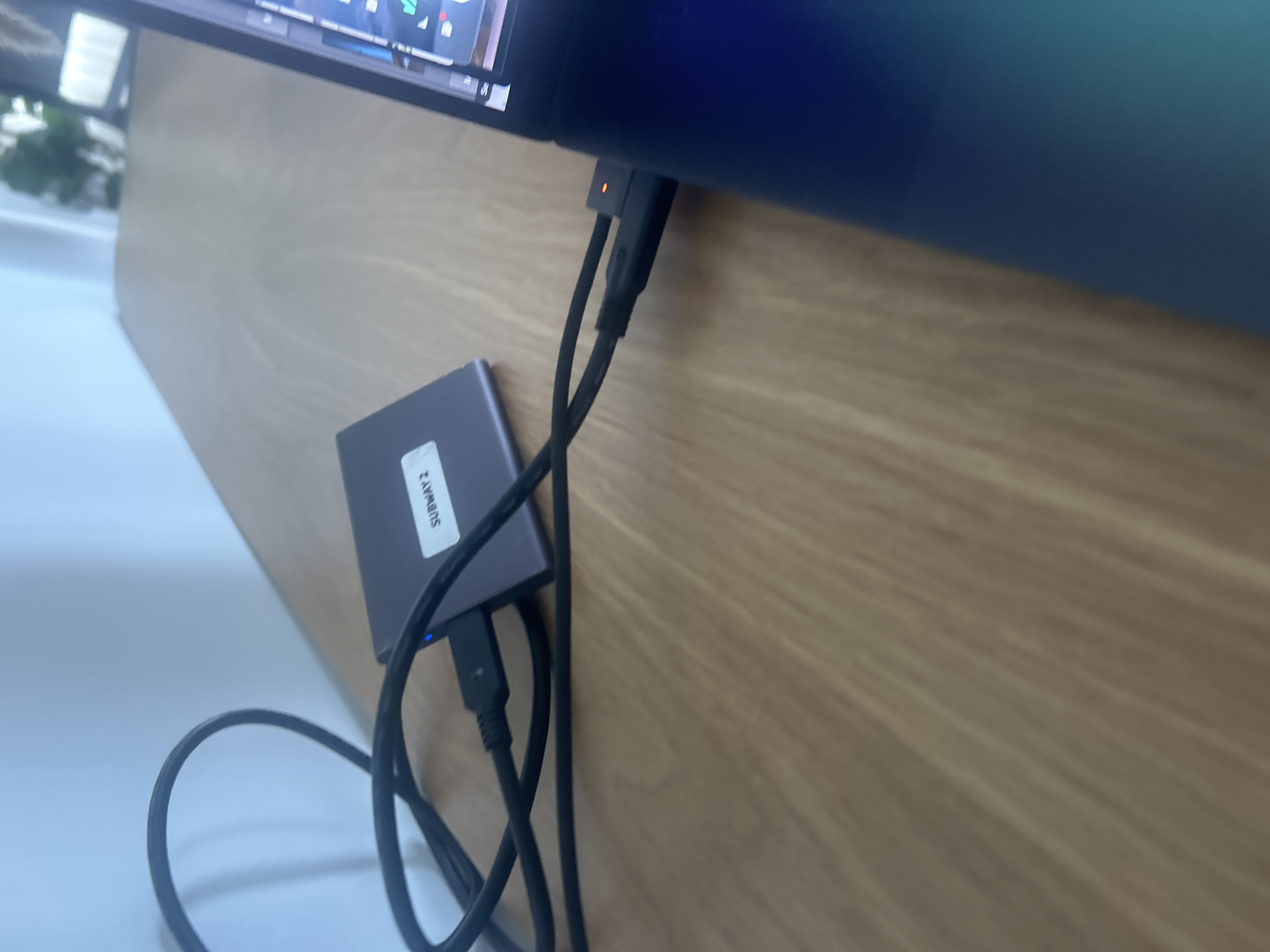 The image shows an external hard drive labeled "SUBWAY 2" connected to a computer. The computer is positioned on a desk with a wooden-grain surface. Two USB cables are plugged into the hard drive and the computer. The hard drive has a small blue light illuminated, indicating it's powered on. Part of the computer screen is visible, along with a glimpse of a white surface and greenery in the background. There is no information within the picture that indicates the location of Miami, United States of America.