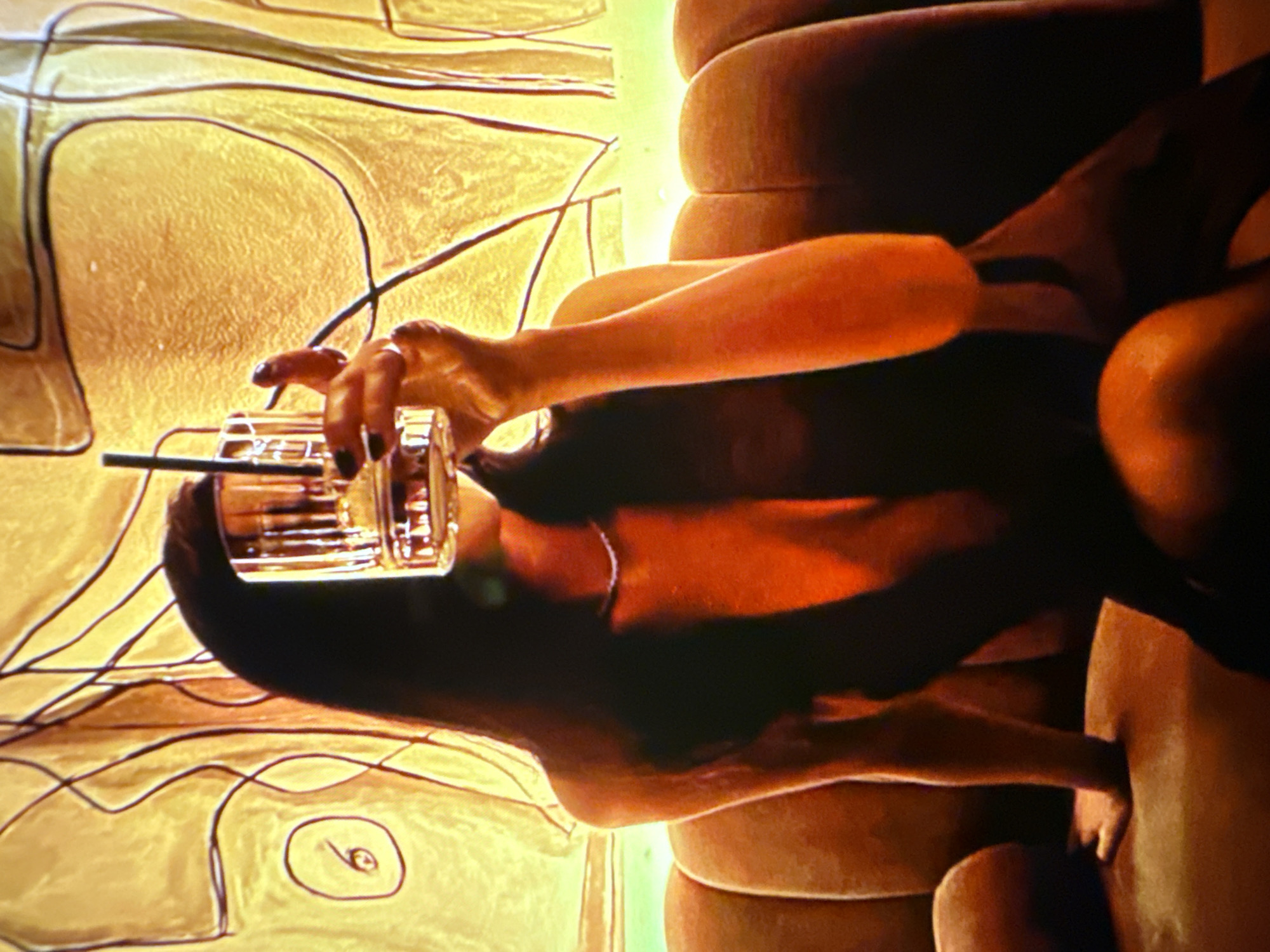 A person with dark hair sits on a tufted orange sofa in front of an abstract yellow and brown wall art. They are holding a glass with a dark straw in their right hand up to their face, partially obscuring their face. The person is wearing a dark, low-cut garment and a thin, sparkly necklace. The background wall art is abstract with flowing lines and a circular shape containing a stylized '6'. The lighting is warm and dramatic, with oranges and yellows dominating the scene.