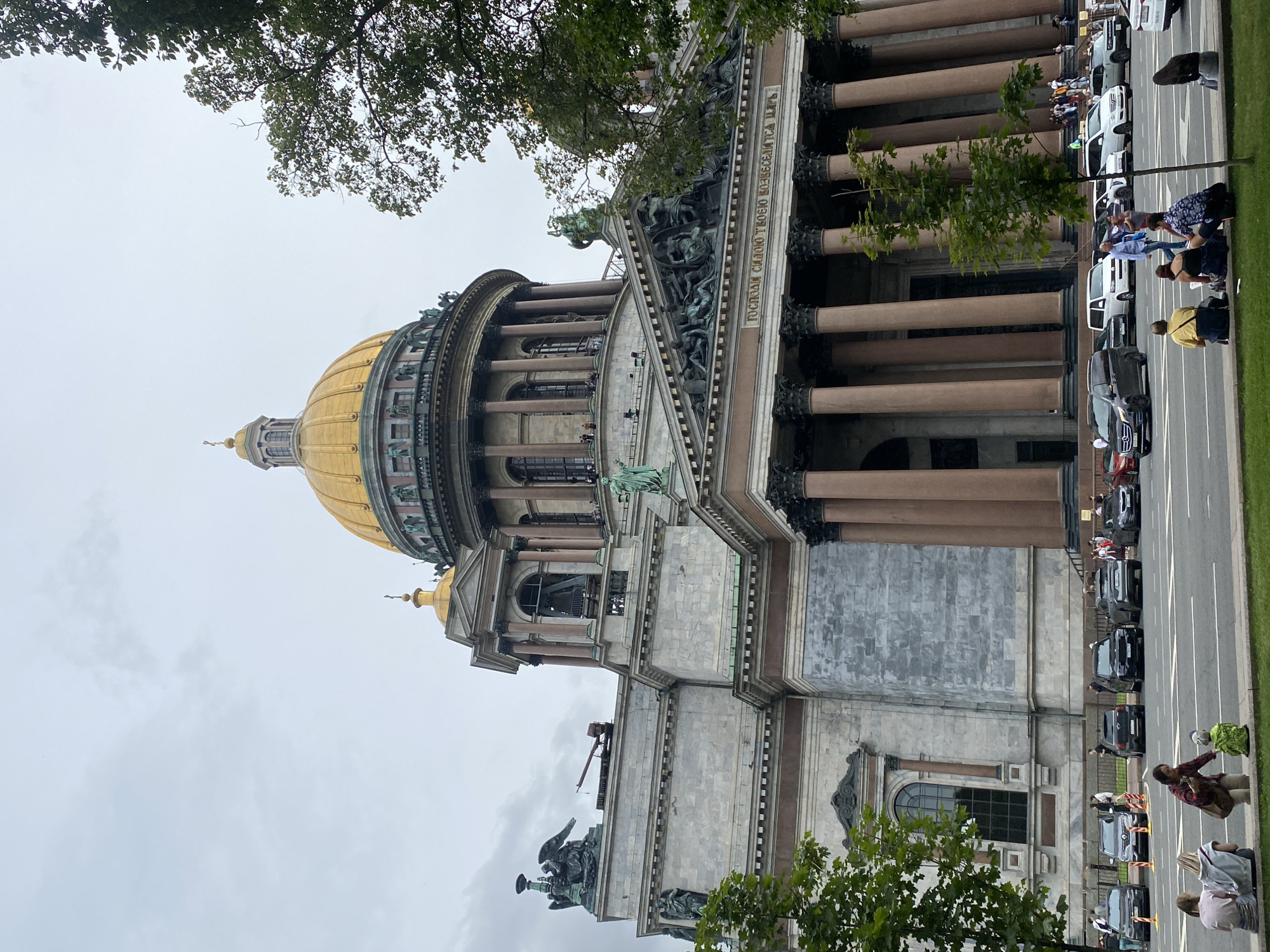 The image shows a low-angle, full-shot view of Saint Isaac's Cathedral in Saint Petersburg, Russia. The cathedral is a massive neoclassical building with a prominent golden dome. The facade features numerous columns, statues, and intricate carvings. In the foreground, a tree partially obscures the view. A street runs along the front of the cathedral, with several cars parked and driving on it. A small group of people are sitting on a grassy area in the foreground, some looking towards the cathedral. The sky is overcast.