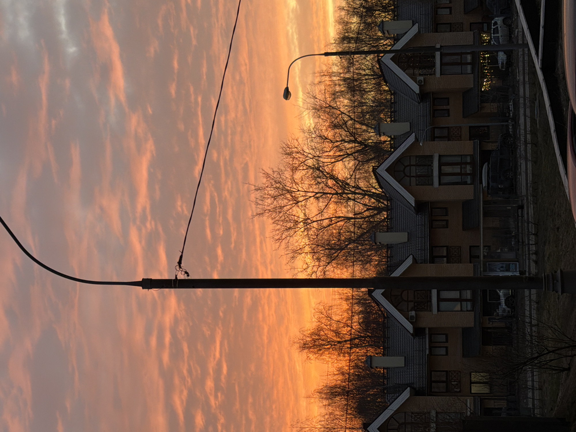 The image depicts a residential street in Khimki, Russia, during either sunrise or sunset. The sky exhibits vibrant orange and red hues with scattered horizontal cloud formations. In the immediate foreground, the top portions of dark vehicles are partially visible, along with sparse, leafless vegetation and a paved surface. A dark, tall utility pole with attached wires stands prominently in the center-left foreground. Another, smaller street lamp with a curved arm is positioned to the right.
The midground features a row of uniformly styled, two-to-three-story residential buildings. These structures have light-brown brick facades and dark-tiled gabled roofs, which include light-colored trim and chimneys. Numerous windows are visible; some are dark, while others reflect the sky or display faint interior illumination, including what appear to be string lights on one balcony. Dark-colored vehicles are parked on the street in front of the residences. In the background, a dense line of bare deciduous trees is silhouetted against the bright horizon. No individuals are discernible within the frame.