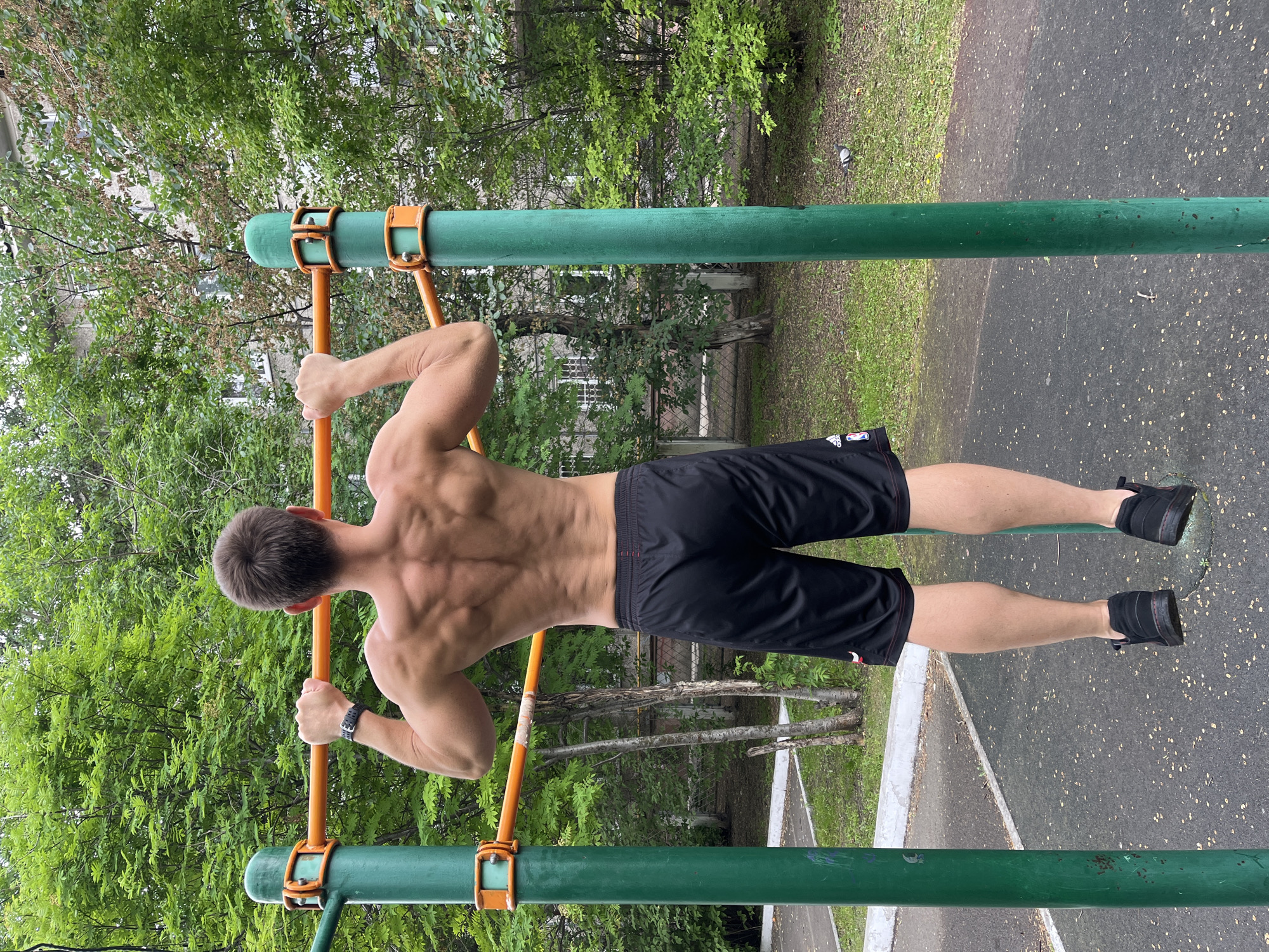 A man is performing a pull-up exercise on an outdoor fitness apparatus in Krasnoyarsk, Russia. The man is shirtless, wearing black athletic shorts and black sneakers. His back is facing the camera, showcasing well-defined musculature. The fitness apparatus consists of two green metal posts supporting an orange horizontal bar. The background features lush green trees, a multi-story building, and a paved ground surface. The image captures a moment of physical exertion in an urban outdoor environment.