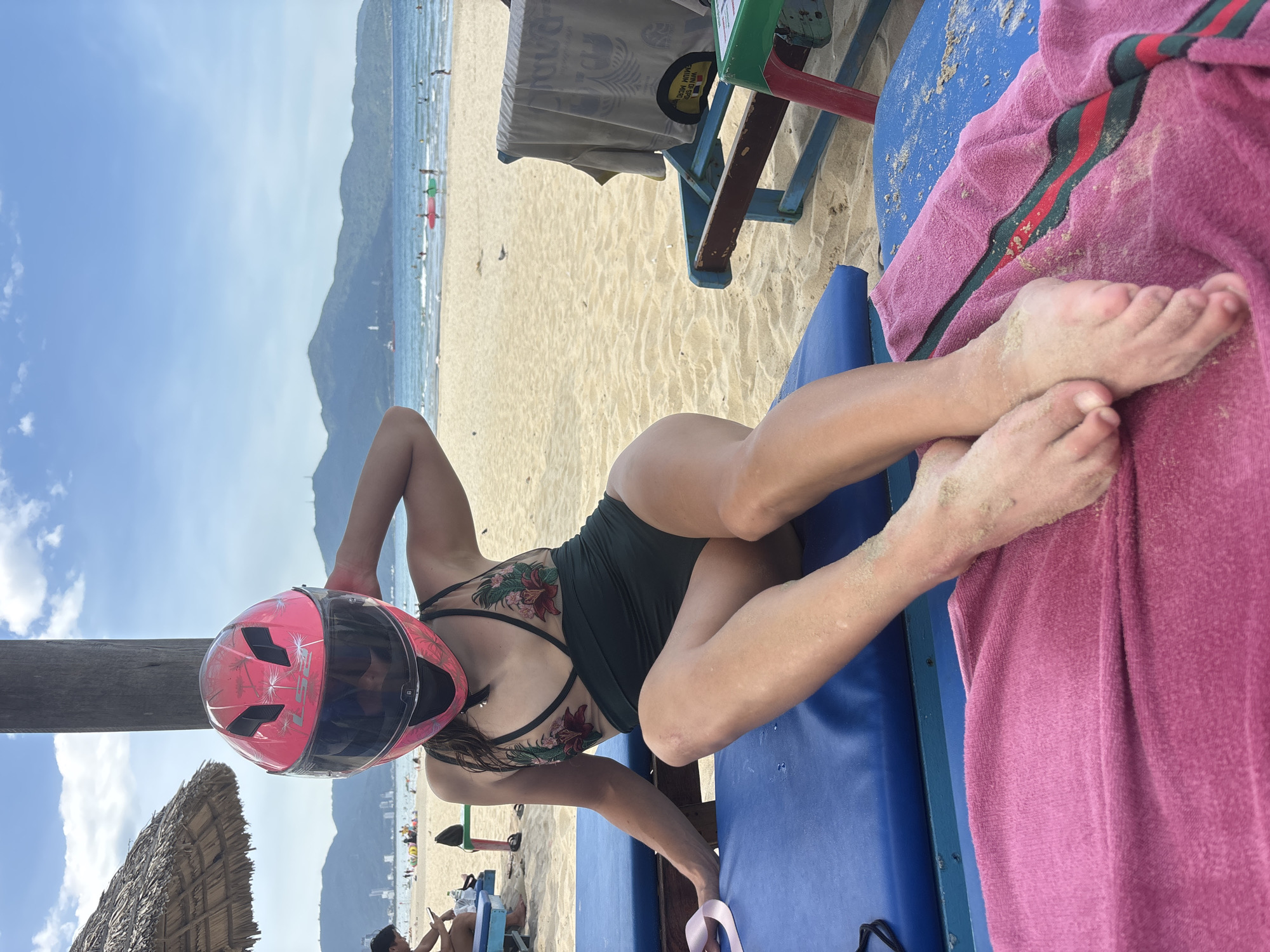 An adult female wearing a pink motorcycle helmet with a floral pattern and a black swimsuit with floral embroidery is sitting on a blue lounge chair on a sandy beach. Her left hand is on the helmet, and her right arm is bent at the elbow, with her hand resting on her head. Her legs are crossed, and her feet are visible at the bottom of the frame, covered in sand. A pink towel is draped over the lounge chair in front of her. In the background, the ocean, other beachgoers, and mountains are visible. The image appears to have been taken on a sunny day. This scene is likely located on a beach in the city of Đà Nẵng, Vietnam, known for its numerous beaches and coastal resorts.