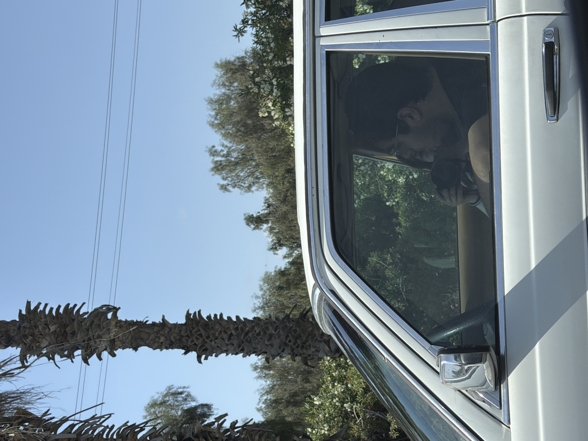 A man with glasses sits in the driver's seat of a white car, holding a black object. The car appears to be on a road with trees, palm trees, and power lines visible outside the car window. The location is the Shafir Regional Council, Israel.