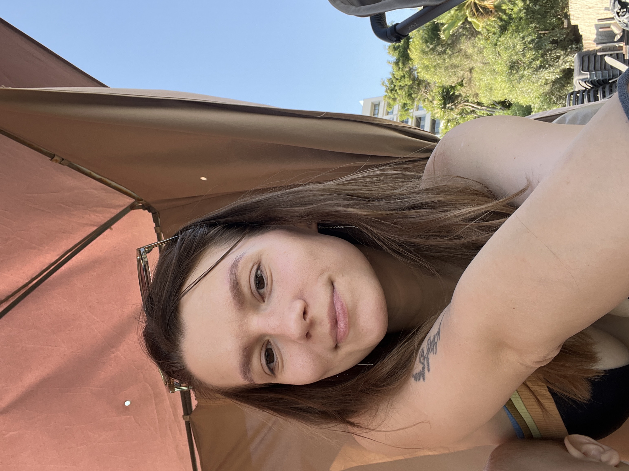 A young Caucasian woman with light brown hair is pictured from the chest up, smiling faintly. She is wearing a dark top with colorful stripes on the shoulder strap. Her left arm is visible, resting on her chest, and a tattoo of branches and flowers is visible on her upper arm. She is seated under a light brown umbrella. The background shows a clear blue sky, lush green trees, a white building, and stacked lounge chairs, suggesting an outdoor setting like a beach resort. The image appears to be taken in Agios Tychona Community, Cyprus, likely during daylight.