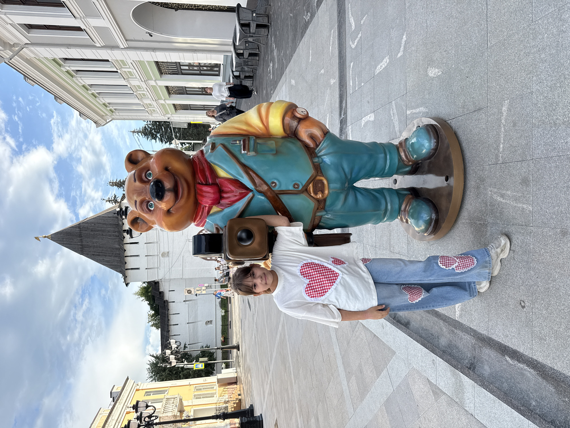 A young girl stands next to a large statue of a bear holding a camera. The girl is wearing a white t-shirt with red and white checkered hearts on it and blue jeans with heart patches. The bear statue is wearing a teal vest, a red scarf, and blue pants. The scene takes place on a city street with buildings in the background and a cloudy sky overhead. The statue is located in Arkhangelsk, Russia, likely near a historical site given the presence of a white tower with a dark roof in the background, which resembles architectural elements of the Arkhangelsk Kremlin. The street is paved with grey stones, and there are lampposts and tables with chairs visible, suggesting a public square or pedestrian area. Two women are walking in the background.