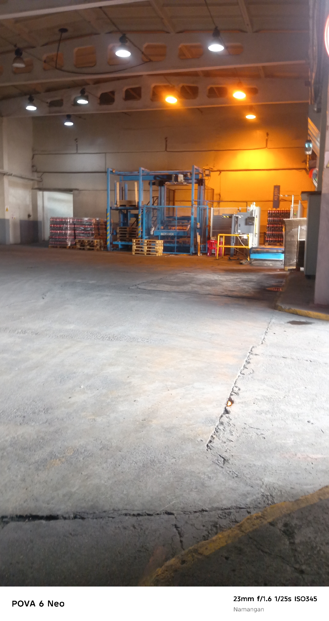The image depicts the interior of a warehouse or factory in Namangan City, Uzbekistan. The scene is characterized by a concrete floor with visible cracks and yellow markings. Industrial equipment, including what appears to be a packaging machine or conveyor system, is present, painted in blue and grey. Stacks of products, possibly bottled beverages due to their red labels and arrangement, are visible on pallets. Overhead, fluorescent lights and orange industrial lamps illuminate the space. The overall environment suggests a functional, operational industrial setting.