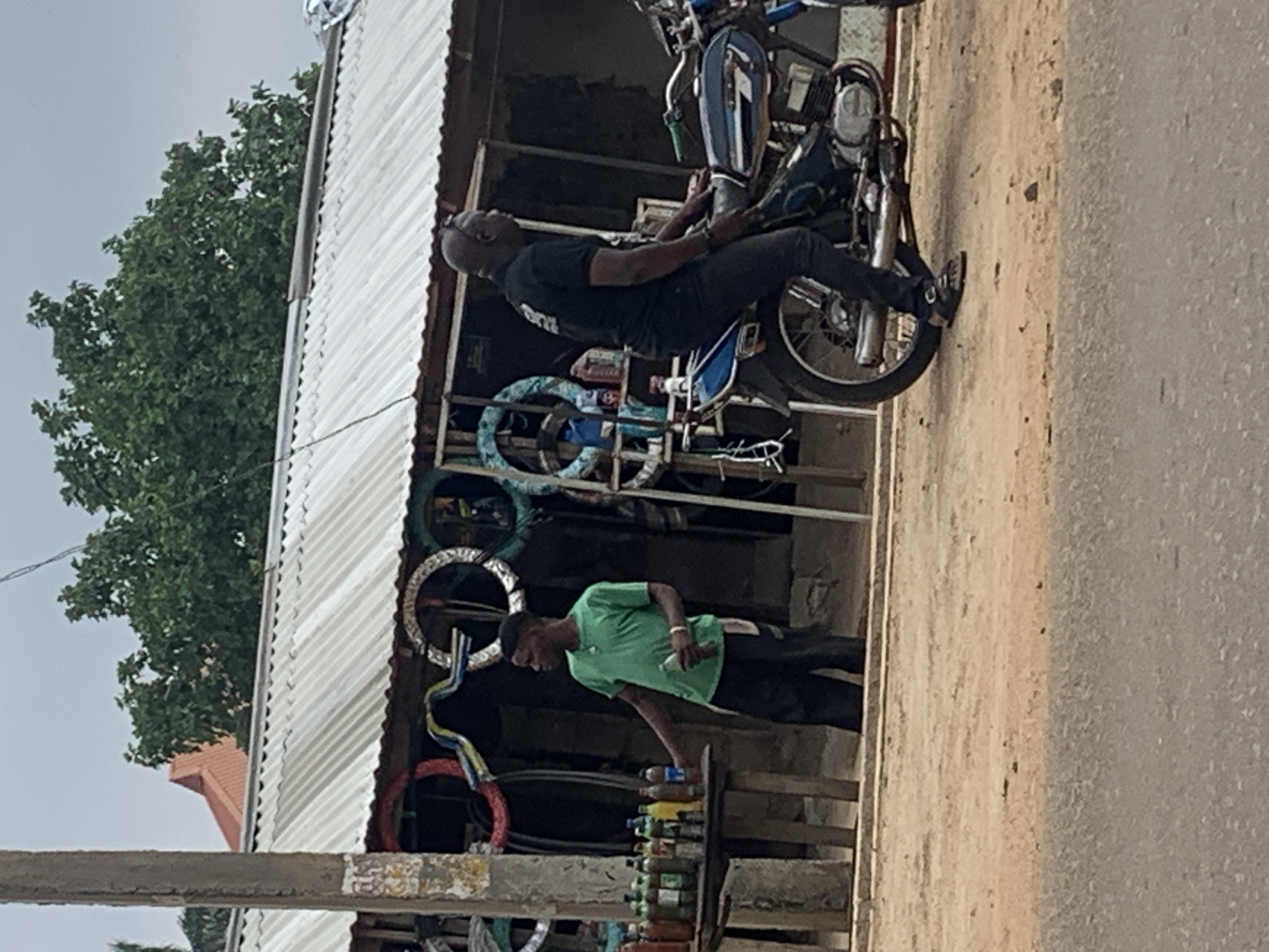 The image captures a casual roadside scene in Maigizo, Nigeria, likely during the daytime under an overcast sky. The setting features a simple, single-story building on the left, characterized by a corrugated metal roof and an open-fronted stall area. A large, leafy green tree is visible behind the building, contrasting with the pale, cloudy sky. The ground on the right appears to be a mix of dirt and rough pavement, suggesting a street or pathway.
Two men are the main subjects. On the left, an older, bald man with dark skin is seated on a motorcycle. He wears a dark t-shirt and dark pants and appears to be looking slightly upwards or ahead, possibly resting or waiting. The motorcycle is a blue and silver model, suggesting a utilitarian vehicle common in the region.
To the right of the motorcycle, a younger man with dark skin stands behind a makeshift wooden counter that is part of the stall. He is dressed in a bright green t-shirt and dark pants and seems to be attending to the items on the counter. The counter is laden with numerous plastic bottles containing various colored liquids, likely beverages or possibly fuel, offered for sale. Behind this vendor, several circular objects, resembling painted tires or wheel rims, are arranged and hanging, adding a touch of color to the otherwise rustic stall.
The scene depicts a typical everyday interaction or moment at a local roadside shop, with one person waiting or resting and another attending to a small business. No discernible text is visible on clothing or signage.