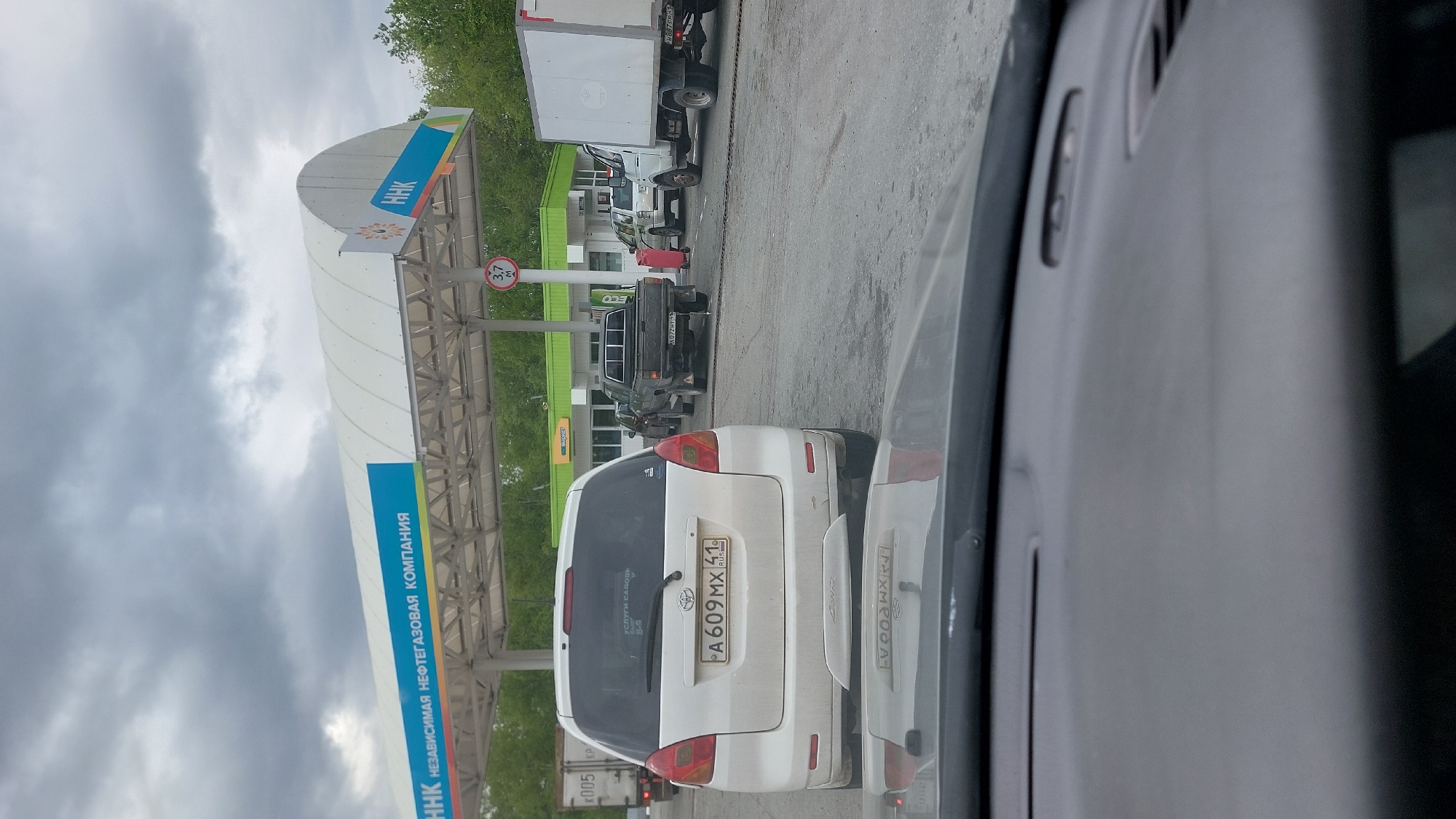 The image shows a queue of vehicles at a gas station in Yelizovo, Russia, under an overcast sky. The gas station canopy features a blue sign with white Cyrillic text that reads "HHK NEFTEGAZOVAYA KOMPANIYA" (HHK Oil and Gas Company). A white Toyota Ractis is in the foreground, facing away from the camera. Its license plate reads "A609MX 41 RUSSIA". Behind it, a dark-colored pickup truck is visible, with license plate "A072HT 41". Further back, a white box truck is parked to the right. A red canister is positioned on the asphalt between the pickup truck and a white SUV. The ground is paved, and trees are visible in the background. The scene suggests a normal day at a Russian gas station, with vehicles lined up to refuel.