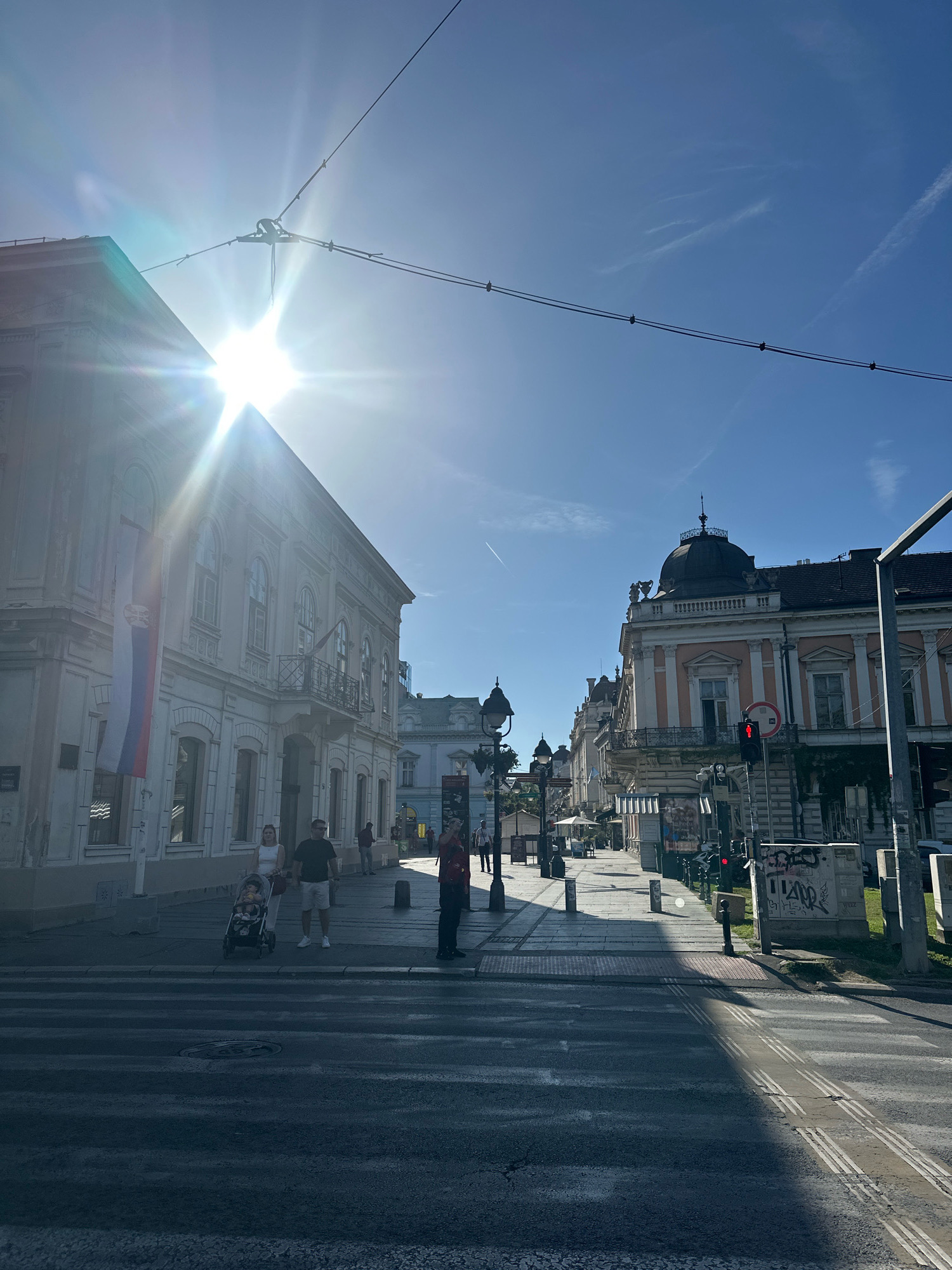 The image depicts a street scene in Belgrade, Serbia, with a prominent sun glare obscuring the sky. On the left, a light-colored building with a balcony features a Serbian flag. Several pedestrians are visible, including a couple pushing a stroller across a crosswalk in the foreground. The street is lined with buildings exhibiting architectural styles characteristic of the city. In the background, more buildings and street elements like lampposts and traffic lights can be seen. The overall environment suggests a sunny day in an urban setting, with a clear blue sky apart from the sun's intensity and some faint contrails.