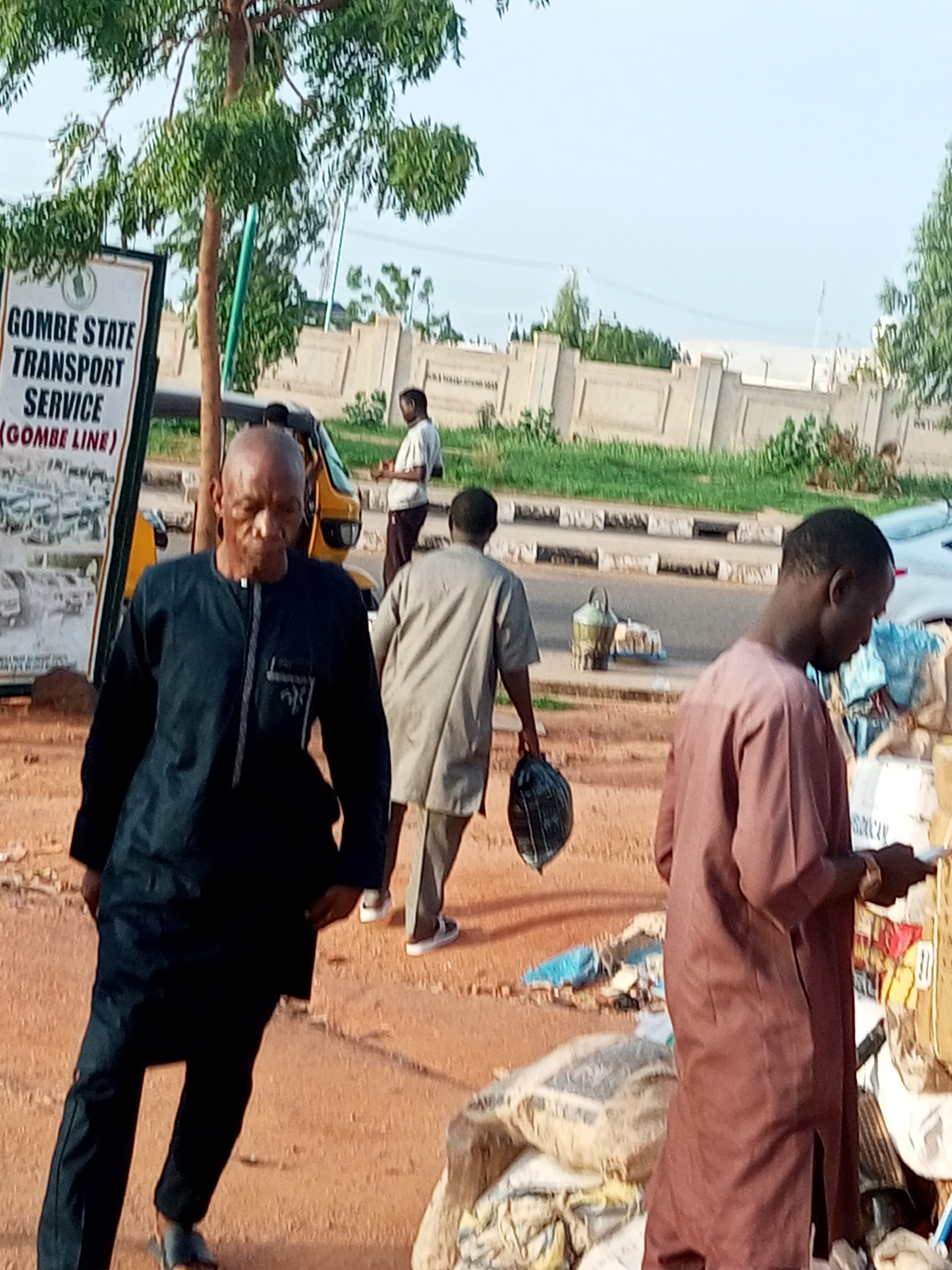 This image captures a street scene in Gombe State, Nigeria, likely near a transportation hub given the prominent sign for "GOMBE STATE TRANSPORT SERVICE (GOMBE LINE)". In the foreground, an elderly man in a dark blue traditional outfit walks towards the viewer, his face visible and his gaze directed downwards. To his right, a younger man in a brown traditional outfit is also in motion, facing away from the viewer. Further in the background, a person in a light-colored outfit stands near a yellow auto-rickshaw, possibly a form of local transportation. The environment appears to be an outdoor setting with a dirt or sandy ground, a road with a curb, and a concrete wall with decorative elements in the mid-ground. Scattered items, including bags and what appear to be market goods, are visible on the right side of the image, suggesting a commercial or roadside vendor area. A tree with green leaves stands to the left of the frame, providing some shade. The overall lighting suggests it is daytime.