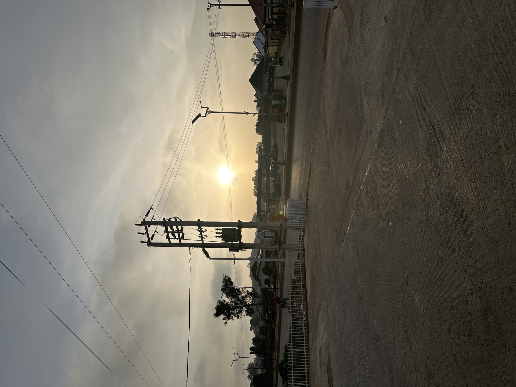 The photograph depicts an outdoor urban scene at sunset in Bukuru, Nigeria. The image is oriented sideways. A paved road dominates the foreground, its surface showing wear. Overhead electrical lines stretch across the sky, connected to a utility pole to the left. A transformer is mounted on the pole. Further down the road, streetlights and buildings are silhouetted against the setting sun. A communication tower stands in the background. Fences and landscaping line the side of the road. A person on a motorcycle is visible near a gated entrance.
