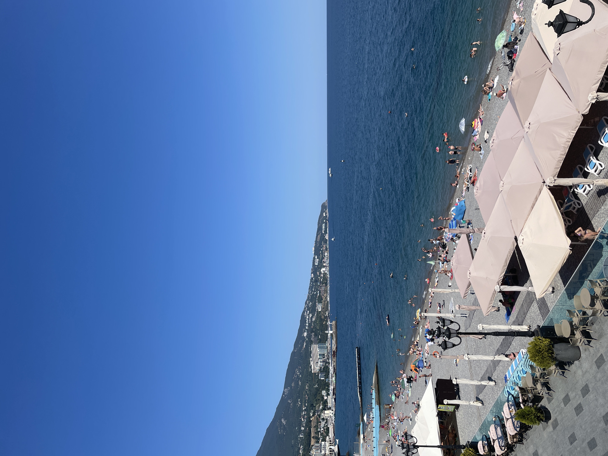 The image depicts a sunny day at a pebble beach resort in Yalta, Ukraine. The foreground features a paved area with outdoor seating, including tables and chairs under tan umbrellas. Beyond this area is a pebble beach populated with numerous sunbathers, many of whom are lying on towels or under umbrellas. People are also seen in the clear blue water, some swimming and others floating on inflatable devices. A pier extends into the sea, and further in the distance, a mountainous coastline with buildings and a lighthouse is visible. The sky is clear and blue.