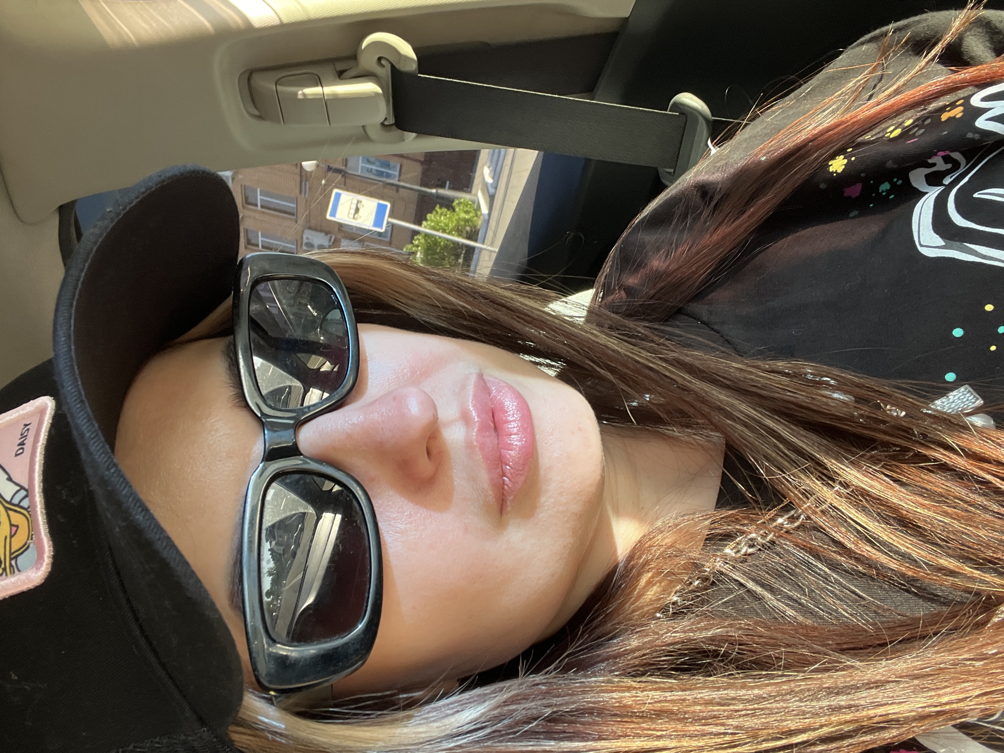 A woman is lying down, presumably in a car, as indicated by the seatbelt visible in the upper portion of the frame. She is wearing a black baseball cap with a "DAISY" patch on it and black-framed sunglasses. Her hair is long and brown with red highlights. She appears to be wearing a black, graphic-printed shirt. Outside the car window, a street sign and buildings are visible, suggesting an urban setting in the city of Unknown, Unknown. The overall lighting suggests it is daytime.