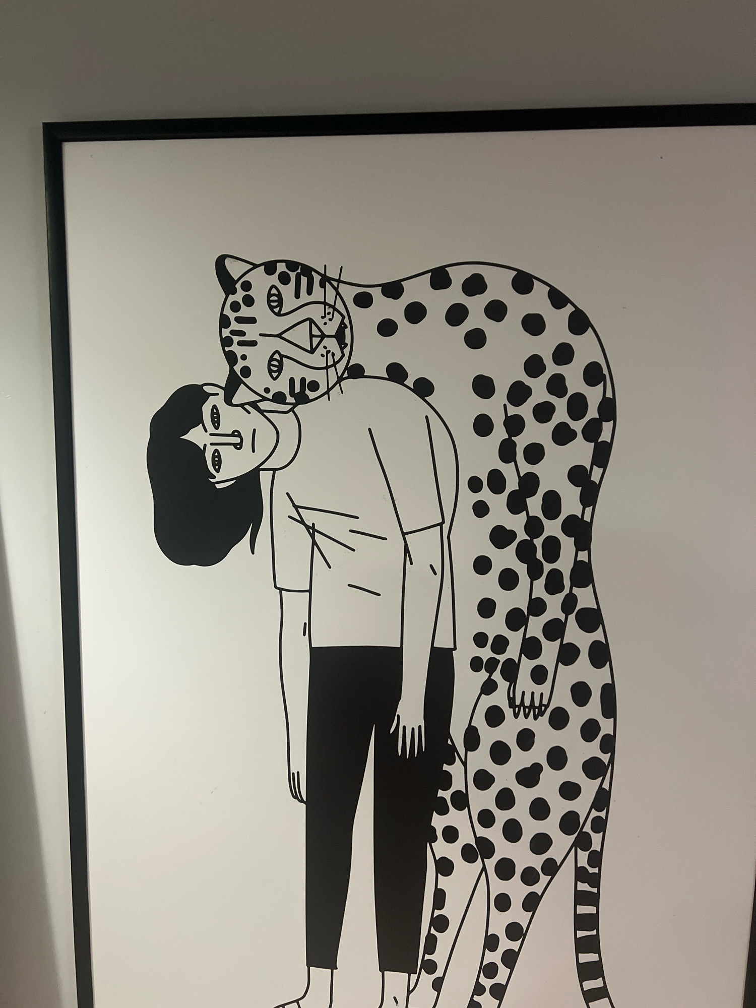 A framed black and white line drawing depicts a person with dark hair, wearing a t-shirt and pants, standing with their head tilted back and resting against a large cheetah. The cheetah's head is positioned over the person's head, and its body extends down the person's back. The drawing is in a minimalist style with bold black outlines and solid black fills for the hair and pants. The cheetah has characteristic spots. The artwork is framed in a black border and appears to be hanging on a light-colored wall. The scene is indoors. There is no indication of the city of El Jadida, Morocco in the image or its context.