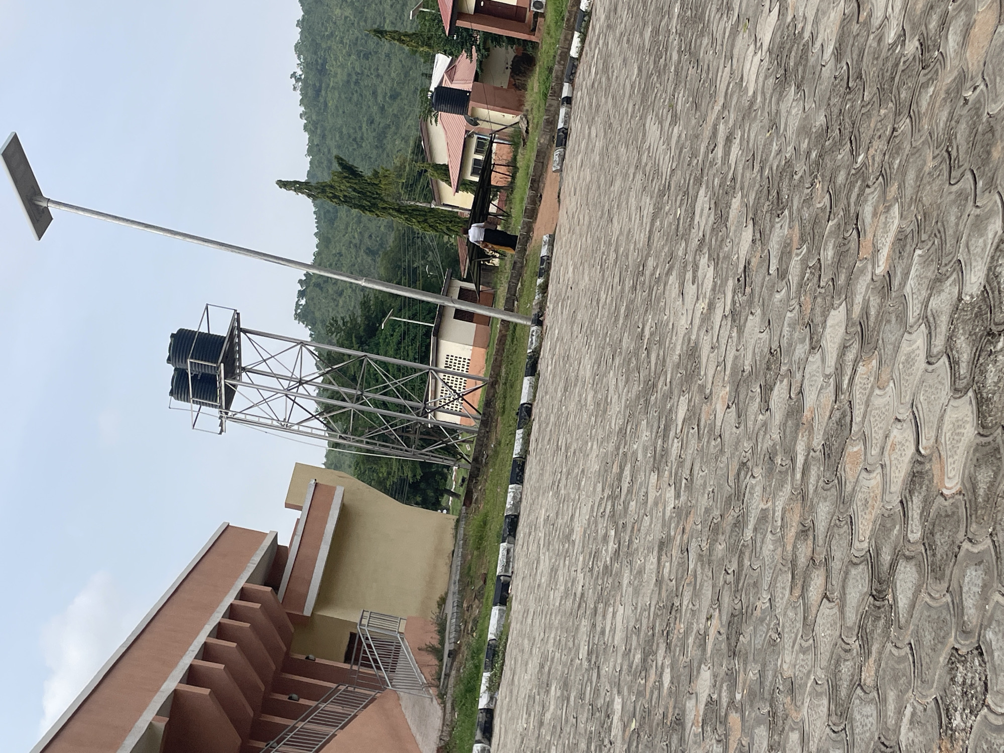 The image depicts an outdoor scene in Lokoja, Nigeria, likely at an educational or administrative facility. In the foreground, a paved area with interlocking hexagonal grey and pinkish concrete blocks extends towards the right and into the distance. A black and white striped curb separates the paved area from a grassy verge.
To the left, a modern building with a pinkish-orange facade and beige lower section features a prominent staircase with metal railings leading to an upper level. A metal water tank on a tall industrial metal framework stands behind this building, and further behind are several smaller, single-story buildings with reddish-brown roofs and beige and white walls, some with visible air conditioning units and windows.
In the middle ground, a lone individual wearing a white shirt and dark trousers walks away from the camera, carrying what appears to be a yellow cloth over their shoulder. They are walking on a dirt path adjacent to the grassy verge.
Dominating the right side of the image is a tall, slender solar-powered street light with a rectangular panel at the top. In the background, lush green hills covered in dense vegetation rise against an overcast sky. The overall impression is one of a functional, somewhat utilitarian campus environment in a hilly, tropical setting.