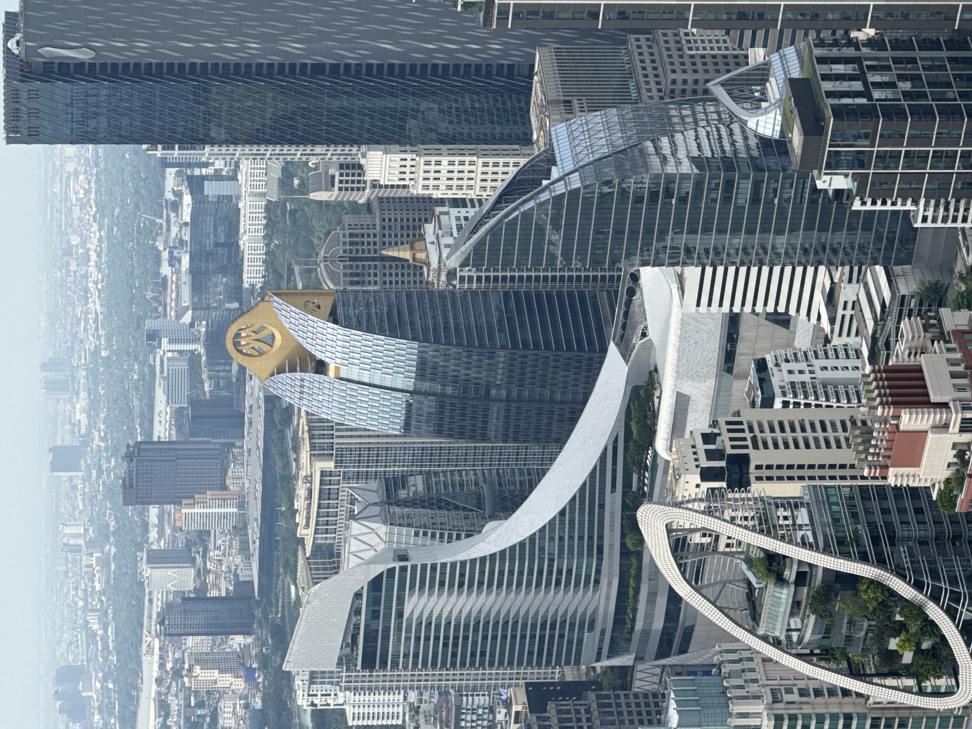 The image presents an aerial view of Bangkok, Thailand, focusing on a cluster of modern skyscrapers. Predominant structures include uniquely designed buildings with curved and angular facades, constructed of glass and steel, and often embellished with gardens and rooftop structures. The city's skyline blends with the cityscape in the background, showing a hazy sky and the tops of other buildings. There is no obvious human activity shown in the picture, and the image seems to prioritize urban architecture.