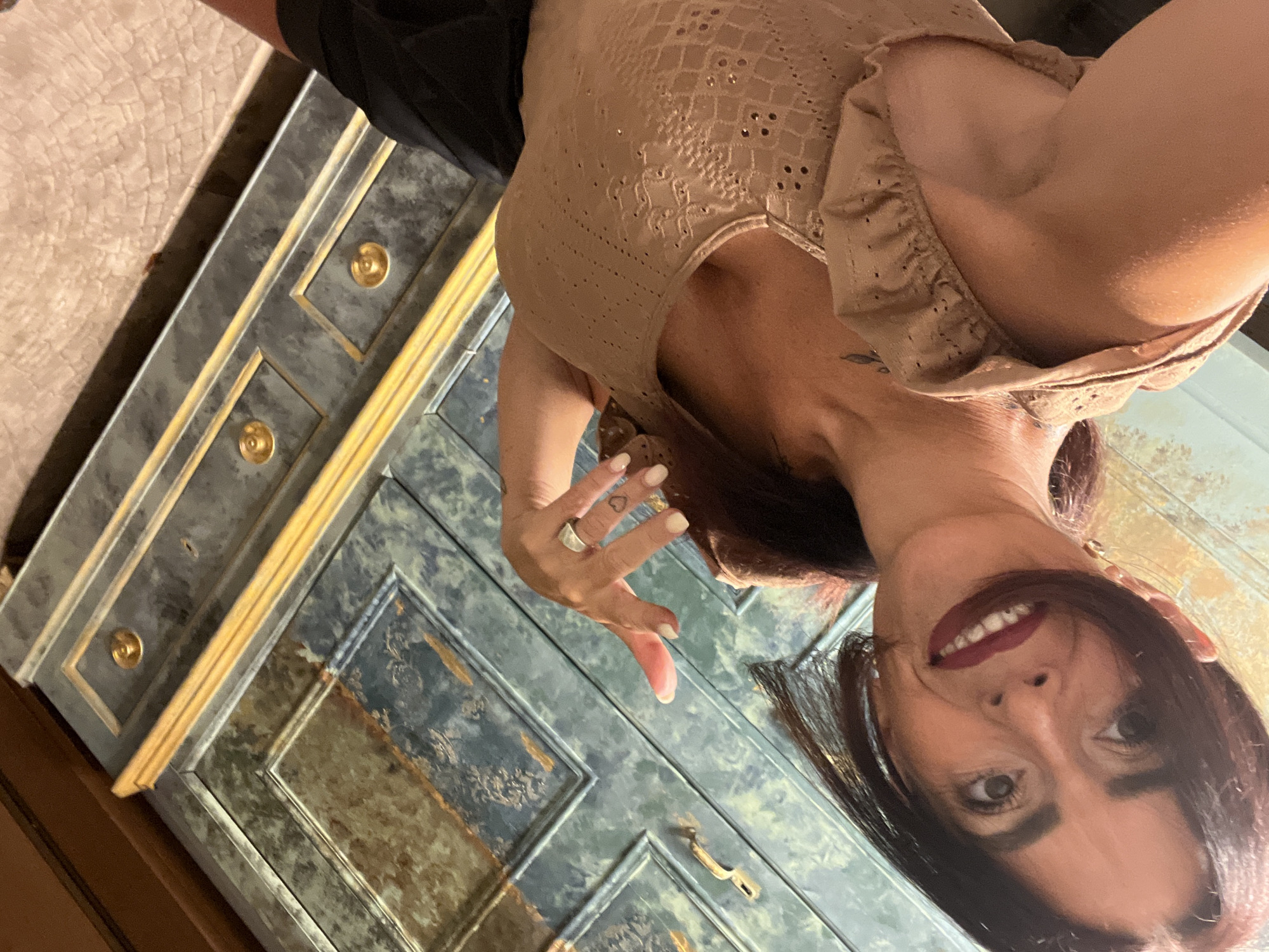 The image shows a woman in a light brown, lace-trimmed tank top and black shorts, taking a selfie in front of a decorative cabinet. She has dark hair and is wearing red lipstick. Her right hand is raised, with fingernails painted white and a ring on her index finger. A faint tattoo is visible on her upper chest. The cabinet has a marbled, blue-green finish with gold trim and drawer pulls. The floor appears to be made of textured tiles. The image was taken in Konz, Germany.