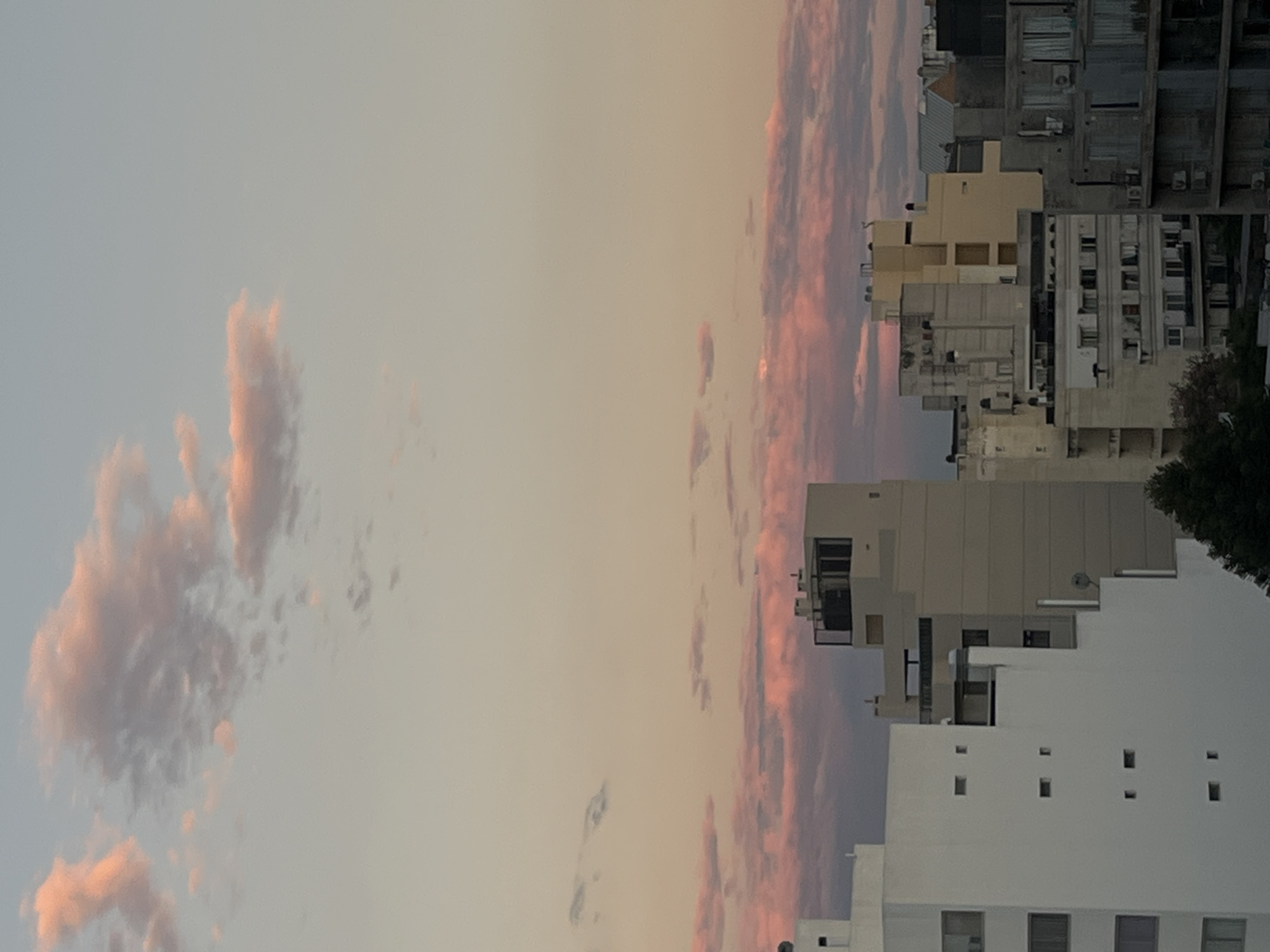 The image shows a cityscape under a sunset sky. Buildings of varying heights and architectural styles dominate the right side of the frame. The sky occupies the majority of the left side, displaying hues of pink, orange, and purple in the clouds. The image seems to be taken during twilight or early evening, as the light is soft and the clouds are illuminated by the setting sun. The location of Cordoba, Argentina is not evident from the image.