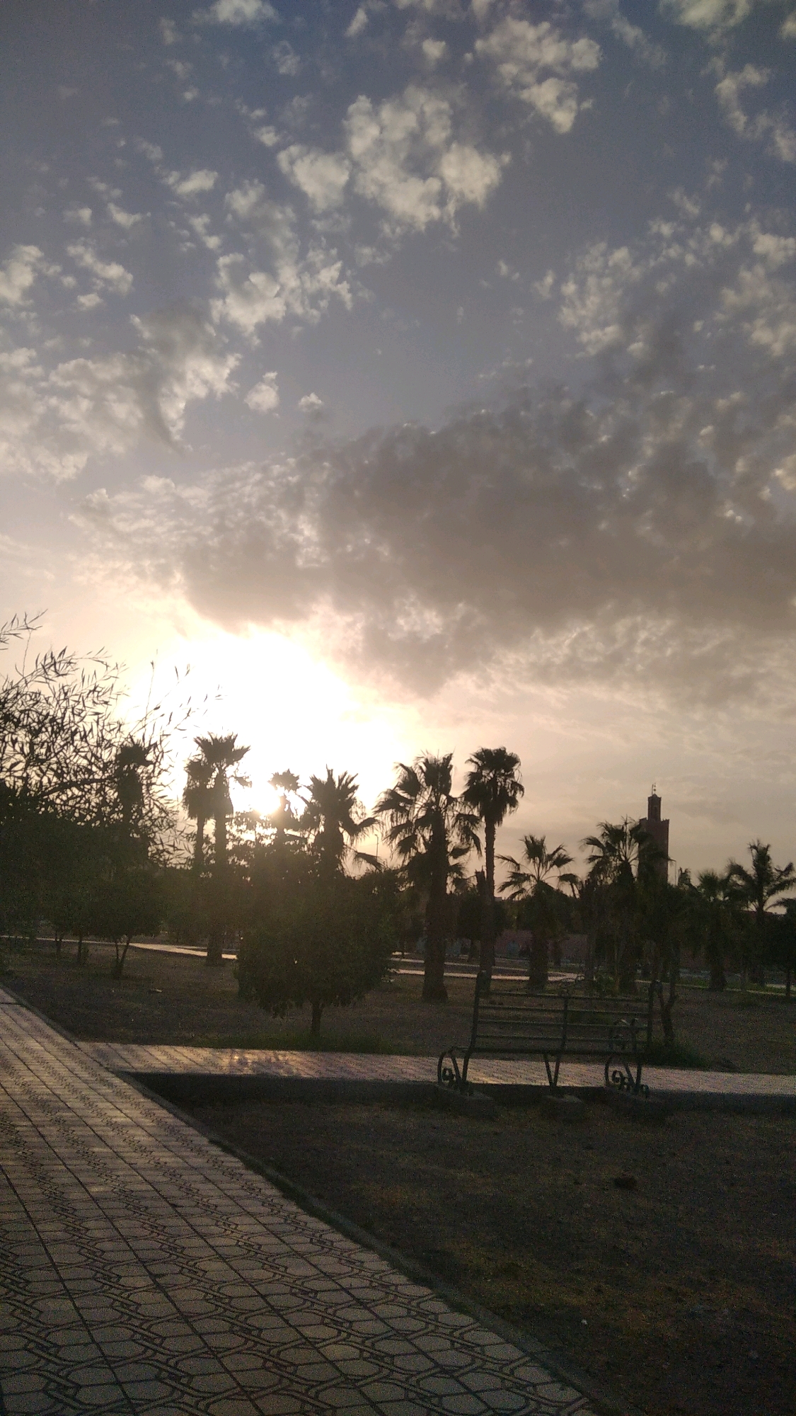 The image depicts a park scene in Marrakech, Morocco, at dusk. The sky is filled with a dramatic display of clouds, with the sun setting behind them, casting a warm, golden glow. Silhouetted palm trees and other vegetation dot the landscape. In the middle ground, a brick pathway with a repeating geometric pattern leads the eye into the park. A park bench is visible to the right of the pathway. In the distance, a minaret of a mosque rises above the trees. The overall impression is one of tranquility and natural beauty as the day comes to a close.