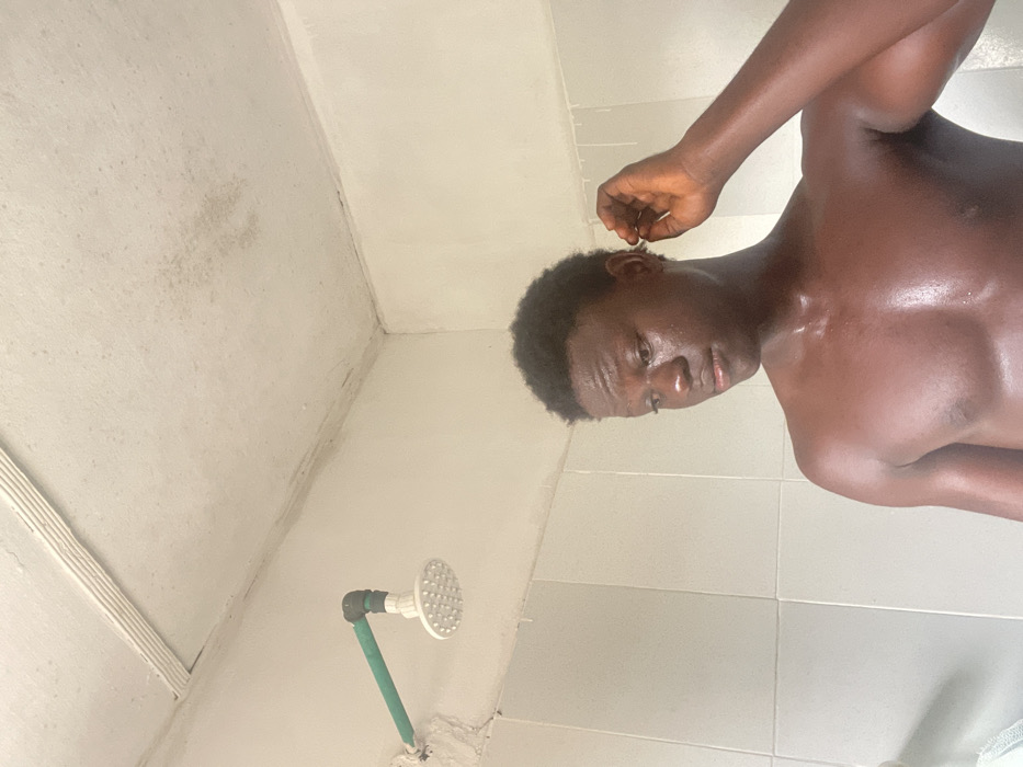 The scene depicts a young adult male in what appears to be a bathroom or shower area in Mosafejaw, Nigeria. The male, with dark skin and short, curly dark hair, is shirtless, revealing his bare chest and shoulders which appear wet or glistening. He is looking downwards and to his right, with his right hand raised near his right ear as if scratching or adjusting something. In the upper left background, a white circular shower head is visible, connected to a green PVC pipe that extends horizontally from a light-colored wall. The wall below the pipe features white tiles. The ceiling above is light-colored and exhibits multiple areas of discoloration and potential moisture damage. The overall environment appears functional but shows signs of wear.