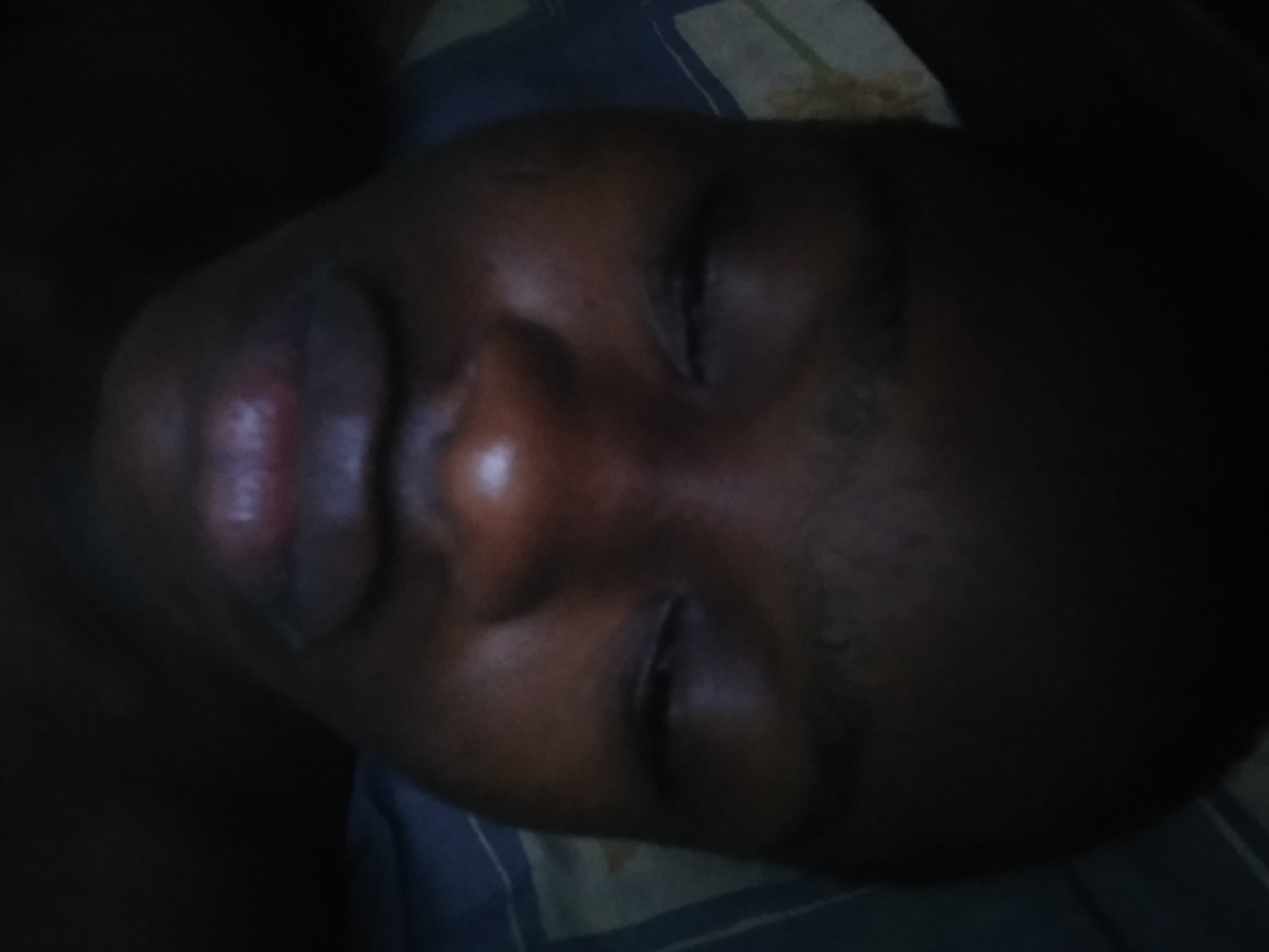The image shows a person lying down with their eyes closed. The person appears to be resting on a bed with a patterned pillow or sheet beneath their head. The lighting is low, making it difficult to discern details, but the person's face is visible, showing their lips, nose, and closed eyelids. The image was taken in a dark or dimly lit room in Shangisha, Nigeria.