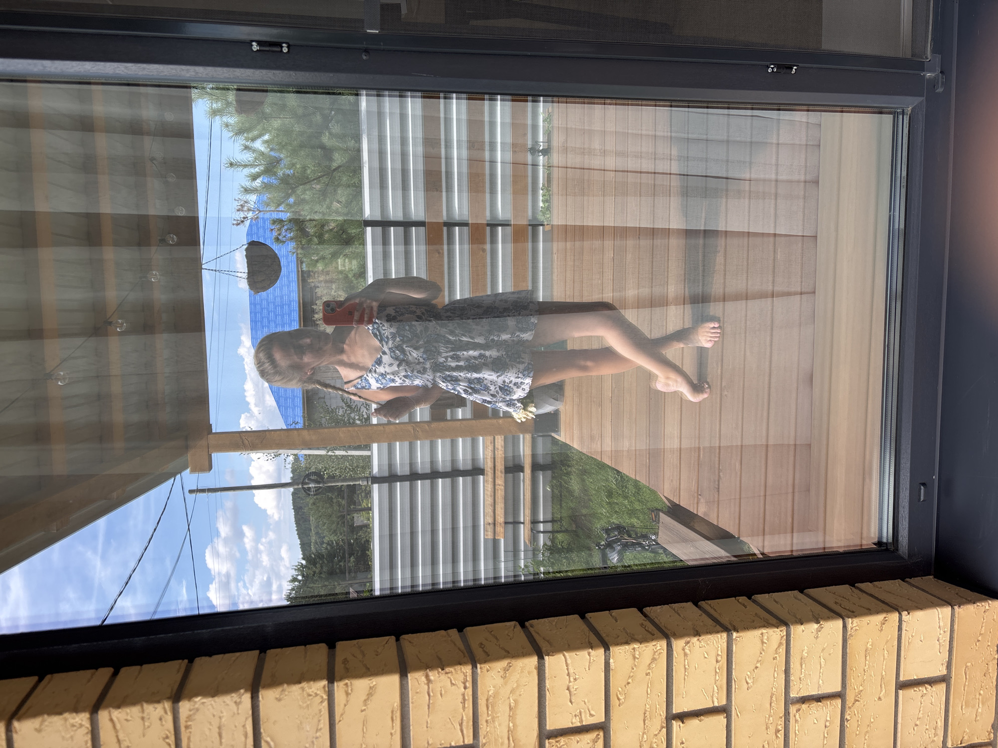 A young woman is reflected in a large glass window. She is standing on a wooden deck, wearing a white and blue floral sundress and holding a red smartphone. Her hair is in two braids. Behind her, a metal fence divides the deck from a grassy area. In the distance, there are trees, hills, and a blue-tiled roof. A wooden bench is visible in the grassy area. The scene appears to be a residential backyard, possibly in Chaikovsky, Russia, given the context. The lighting suggests it is daytime.