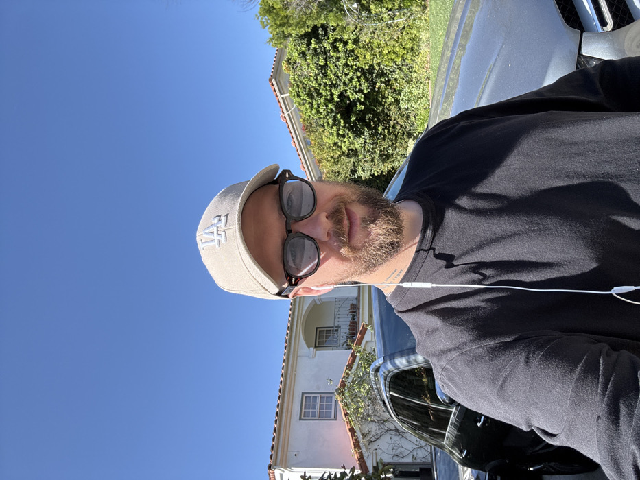 A male subject is positioned centrally in a daylight photograph, appearing to be taking a self-portrait. He wears a beige baseball cap with a white "LA" logo, dark-rimmed sunglasses with tinted grey lenses, a black long-sleeved shirt, and white wired earbuds. The subject has a beard and exhibits a slight smile. Behind him, a black Mercedes-Benz SUV is partially visible, parked in front of a two-story white stucco house featuring a red tile roof and green foliage on its facade. A small balcony with potted plants is visible on the upper floor of the residence. To the subject's right, a dense green hedge lines the background. The sky is clear blue. This scene is located within an upscale residential area of Los Angeles, California, United States.